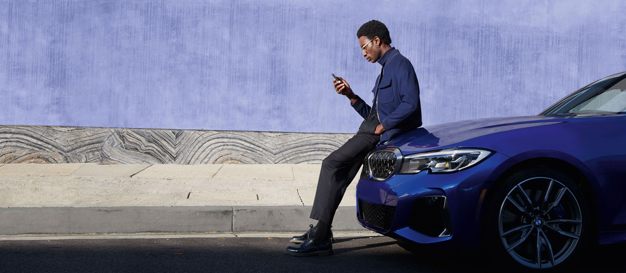 A man leans on the hood of his BMW as he uses his mobile phone.