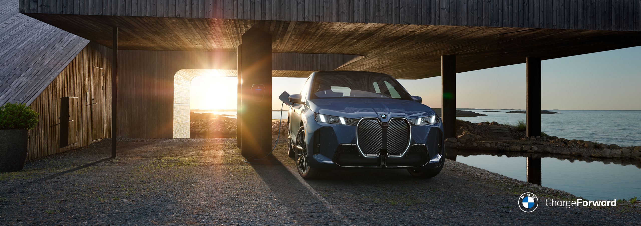 A BMW iX3 charges under a large wooden structure in a coastal area. 