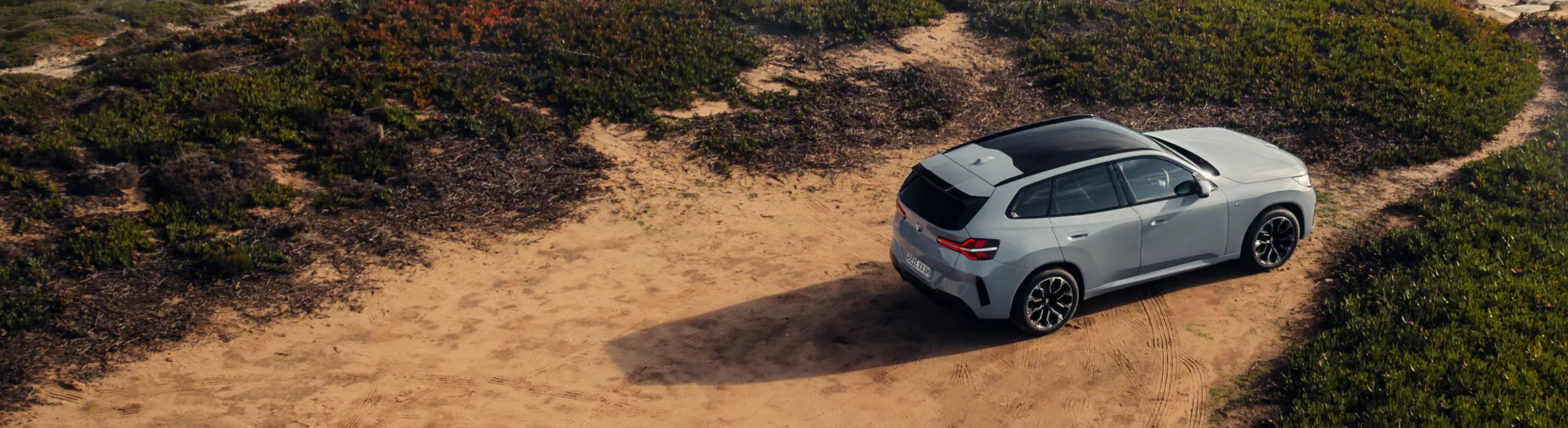  High-angle view of a grey BMW X3 Plug-In Hybrid parked on a dirt path overlooking a rugged coastline with breaking waves