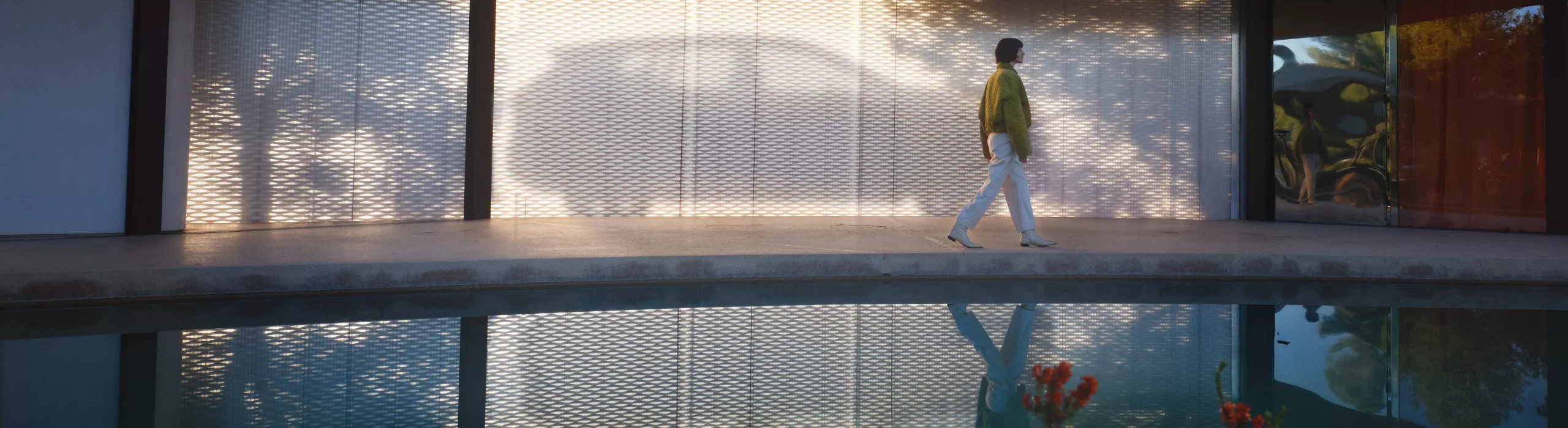 Woman walking beside modern architecture with reflected car silhouette on mesh wall near a tranquil pool