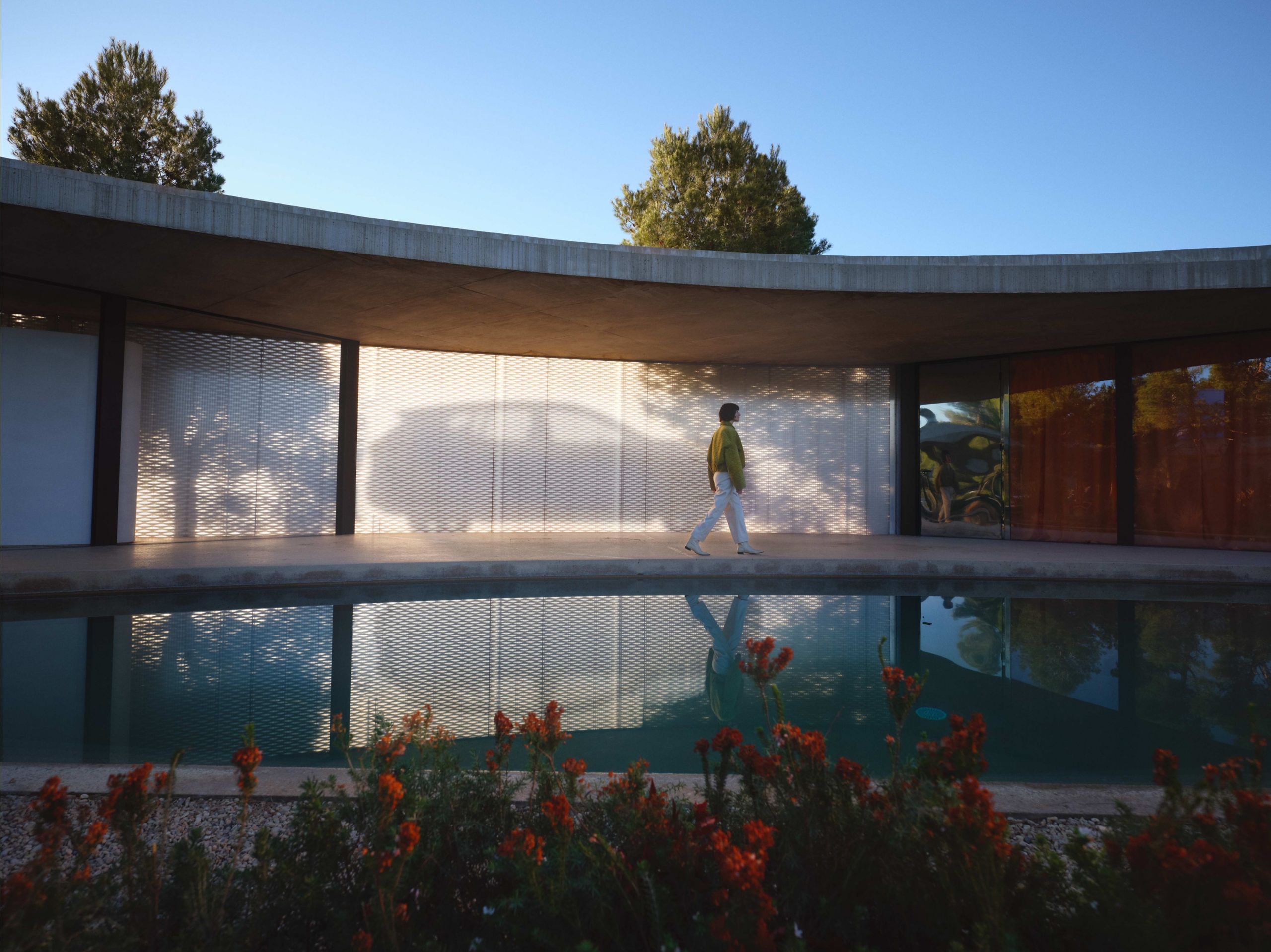 Woman walking beside modern architecture with reflected car silhouette on mesh wall near a tranquil pool