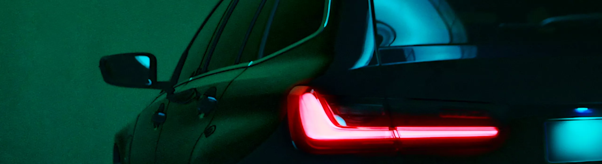 Rear three-quarter view of a dark BMW SUV highlighted in a high-contrast cinematic setting. The glowing red LED L-shaped taillight is sharply in focus, while vibrant neon blue and green ambient lights reflect off the car's body panels and rear window.