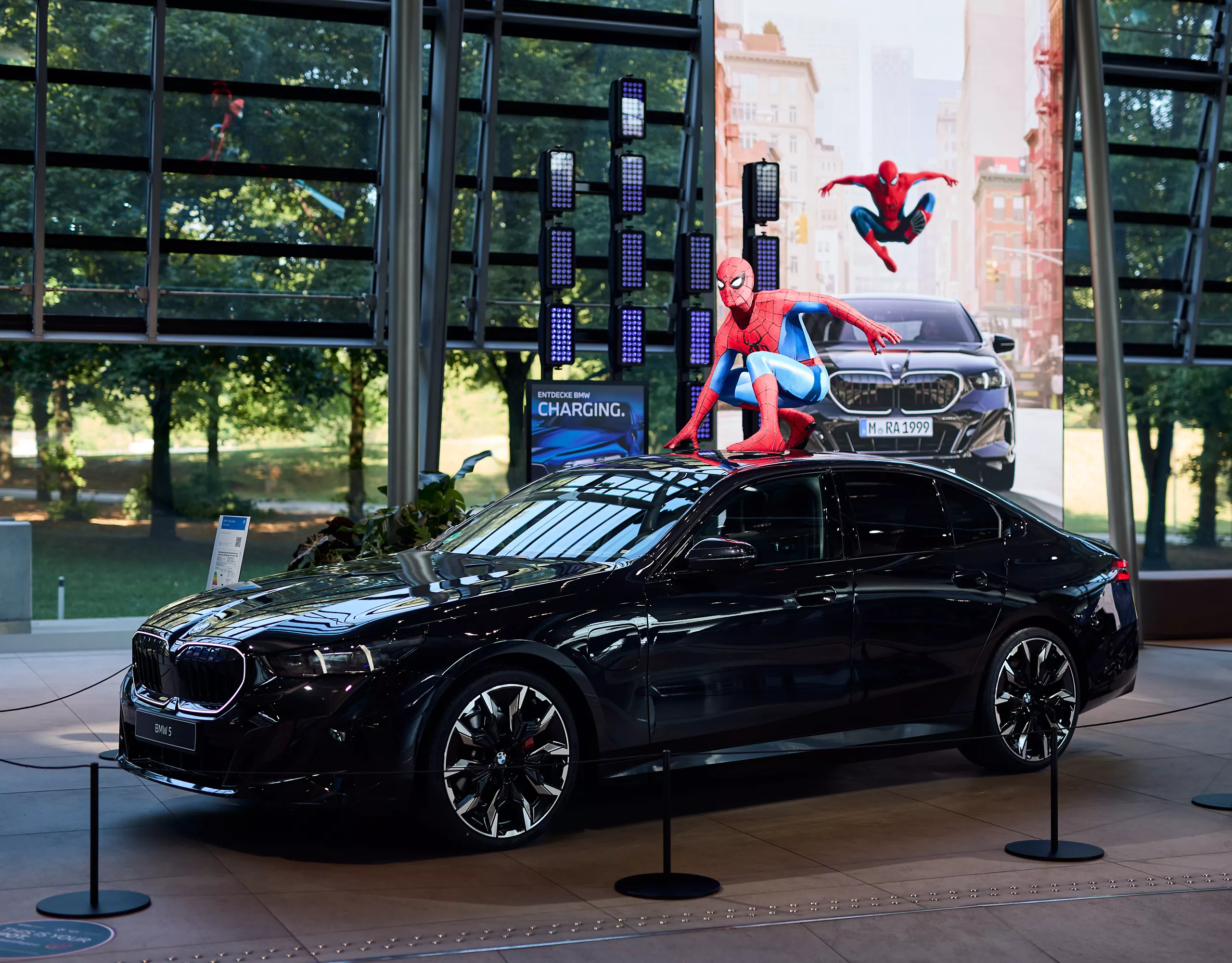 Black BMW 5 sedan displayed at BMW Welt with a Spider-Man figure on the vehicle roof in front of a large Spider-Man themed backdrop.
