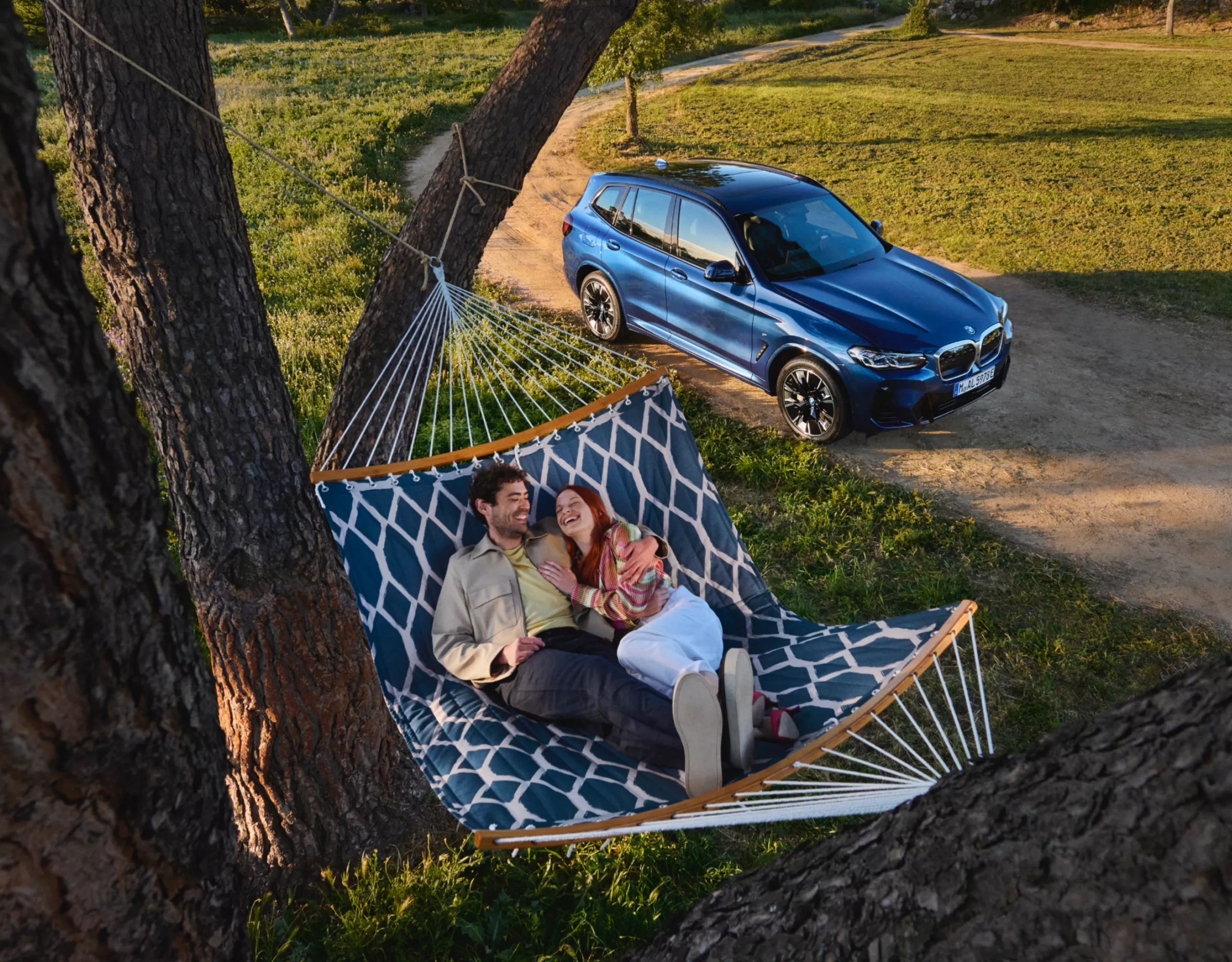 Zwei Personen liegen in einer Hängematte vor einem BMW umgeben von Bäumen