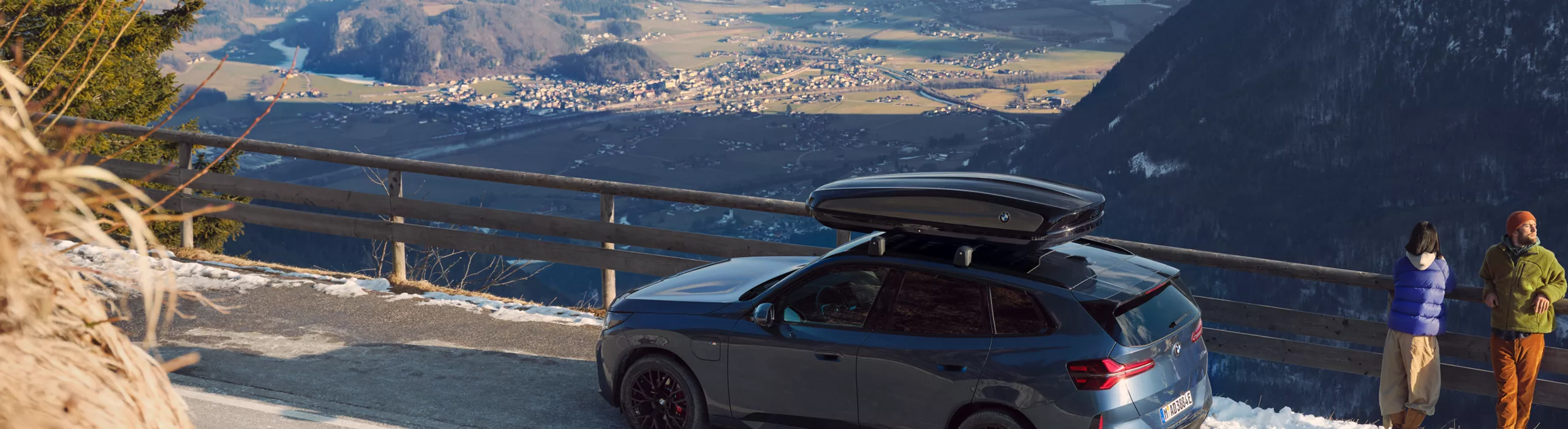 Un BMW con caja de techo aparca en una carretera de montaña mientras dos personas contemplan los valles y las montañas