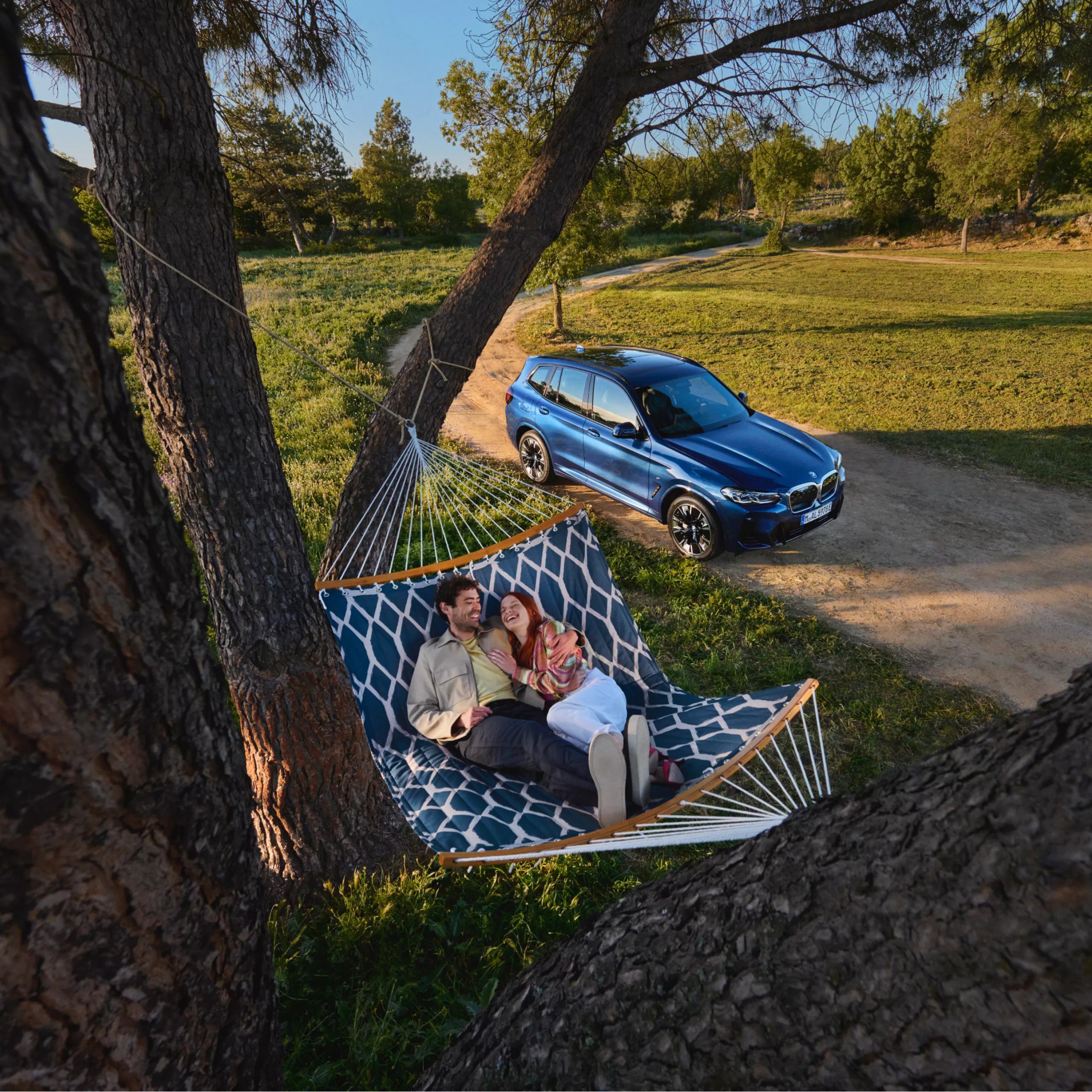 Zwei Personen liegen in einer Hängematte vor einem BMW umgeben von Bäumen