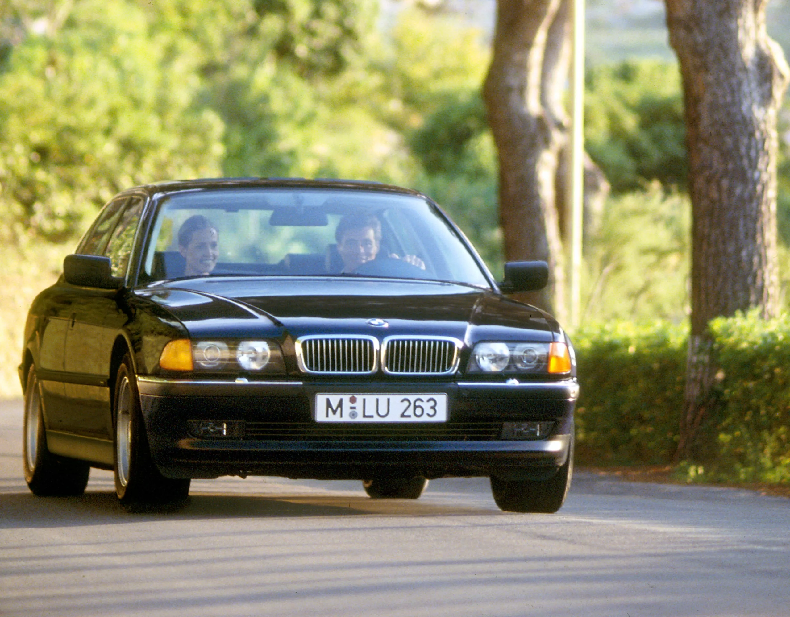 BMW 7er Limousine E38 auf einer Alleenstraße in ländlicher Umgebung im Sommer mit einem Dorf im Hintergrund