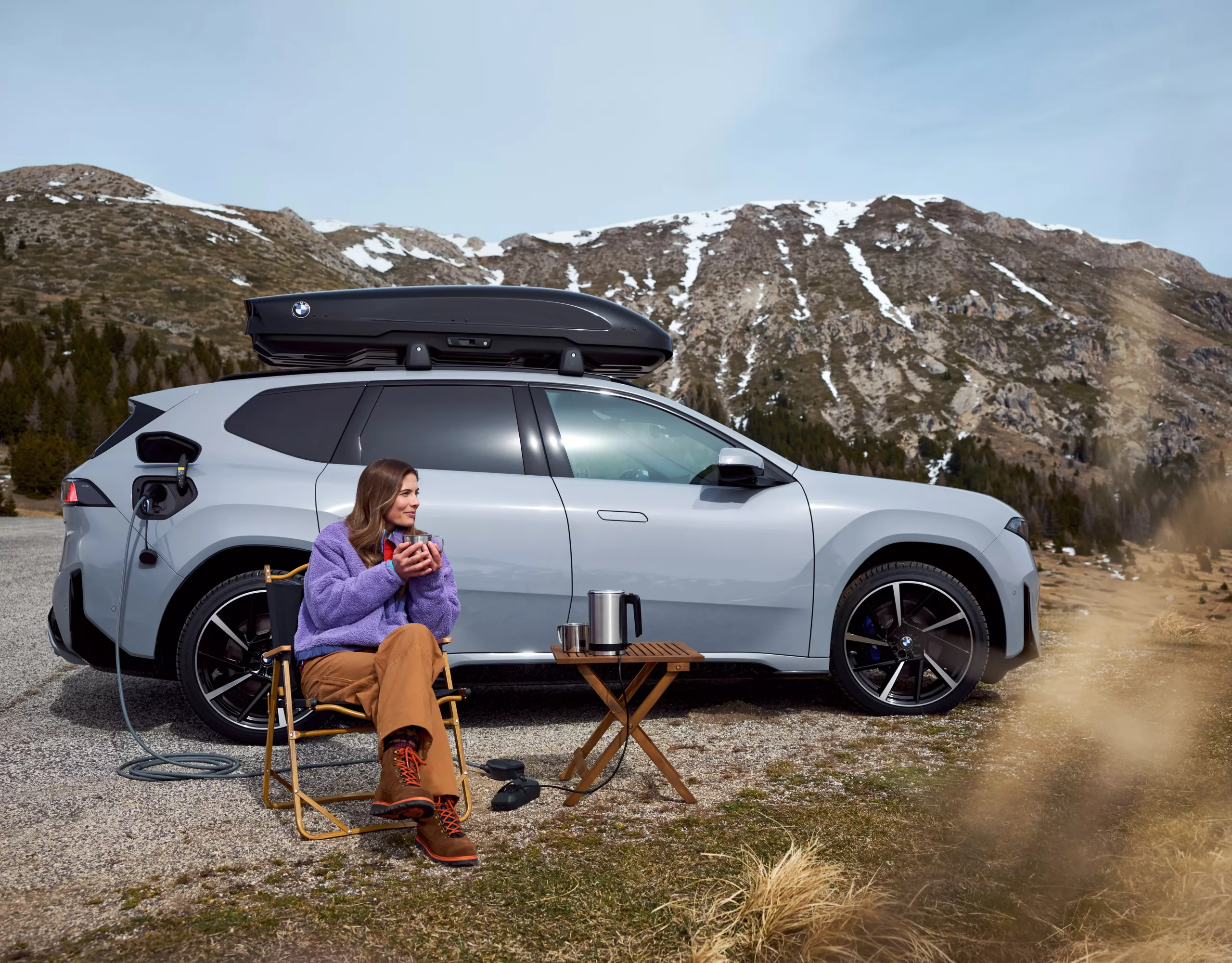 Un SUV électrique BMW en cours de recharge à l'aide d'une borne de recharge mobile ; une personne en camping avec une bouilloire dans un paysage de montagne