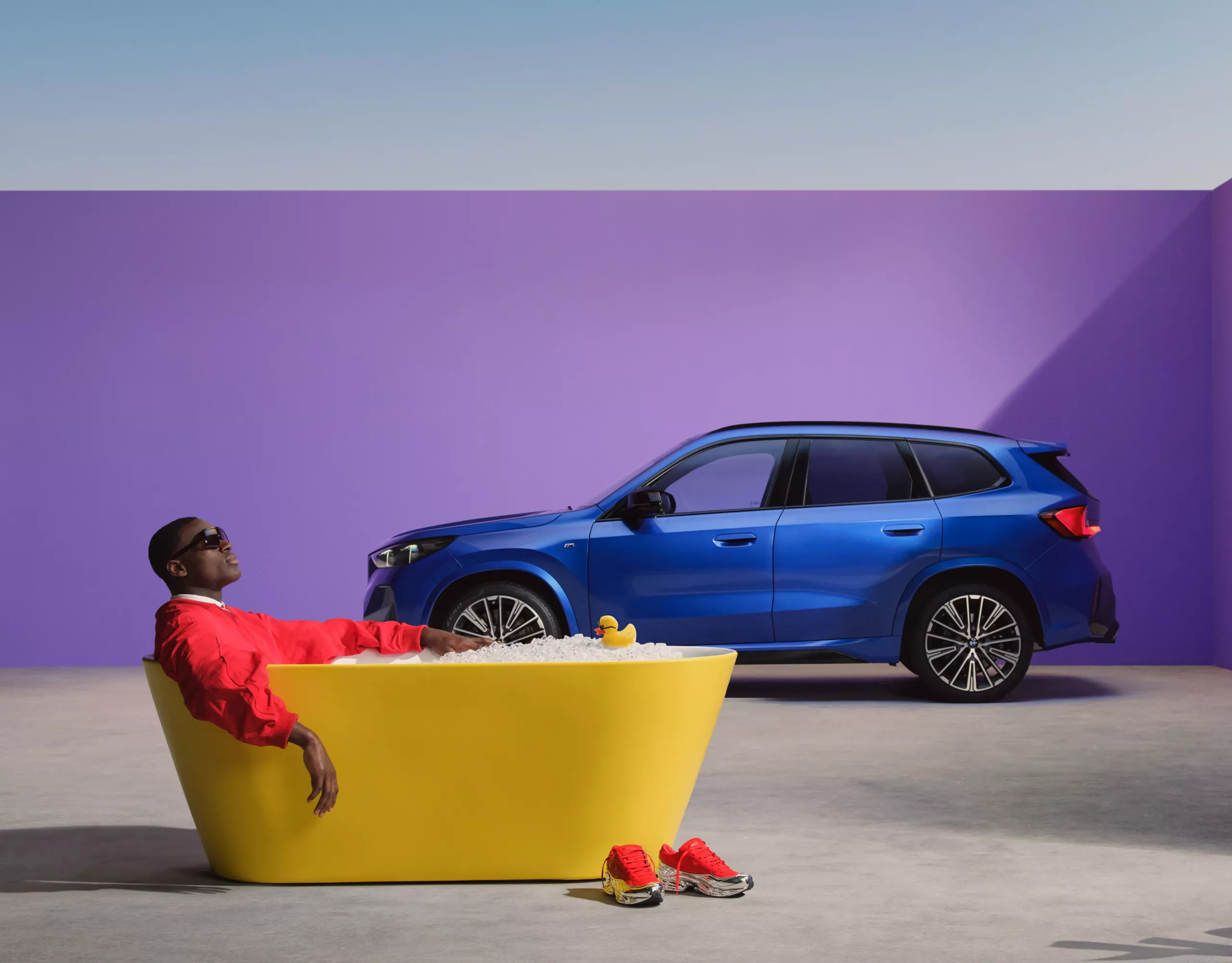 Egy piros ruhás személy egy sárga kádban, lila fal előtt, háttérben egy BMW SUV-val
