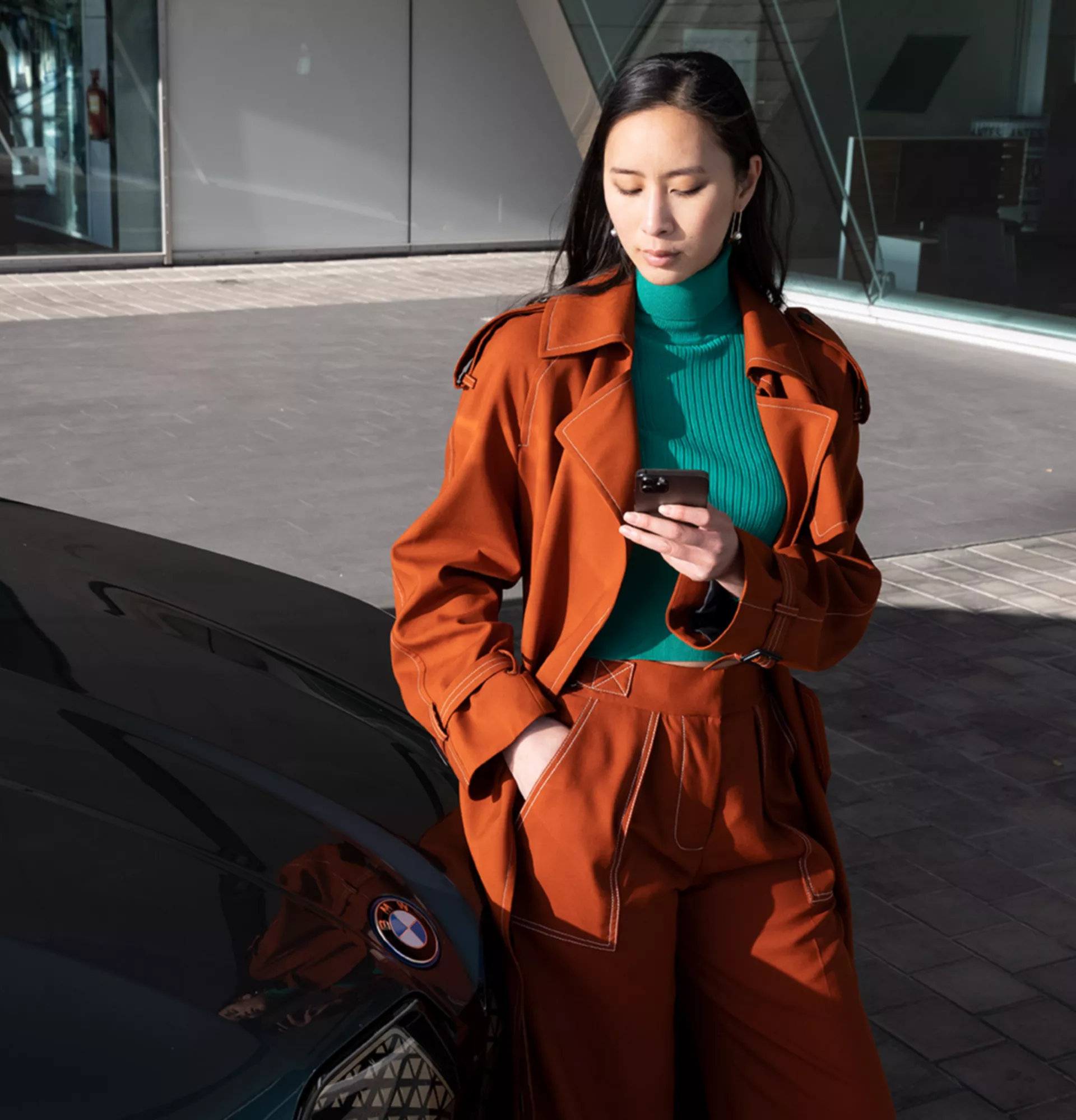 Woman stands next to a BMW in front of office building looking at her smartphone