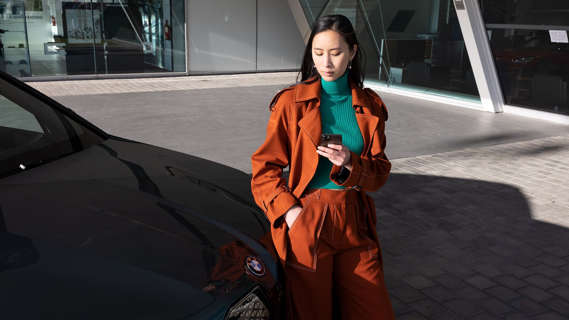 Woman stands next to a BMW in front of office building looking at her smartphone