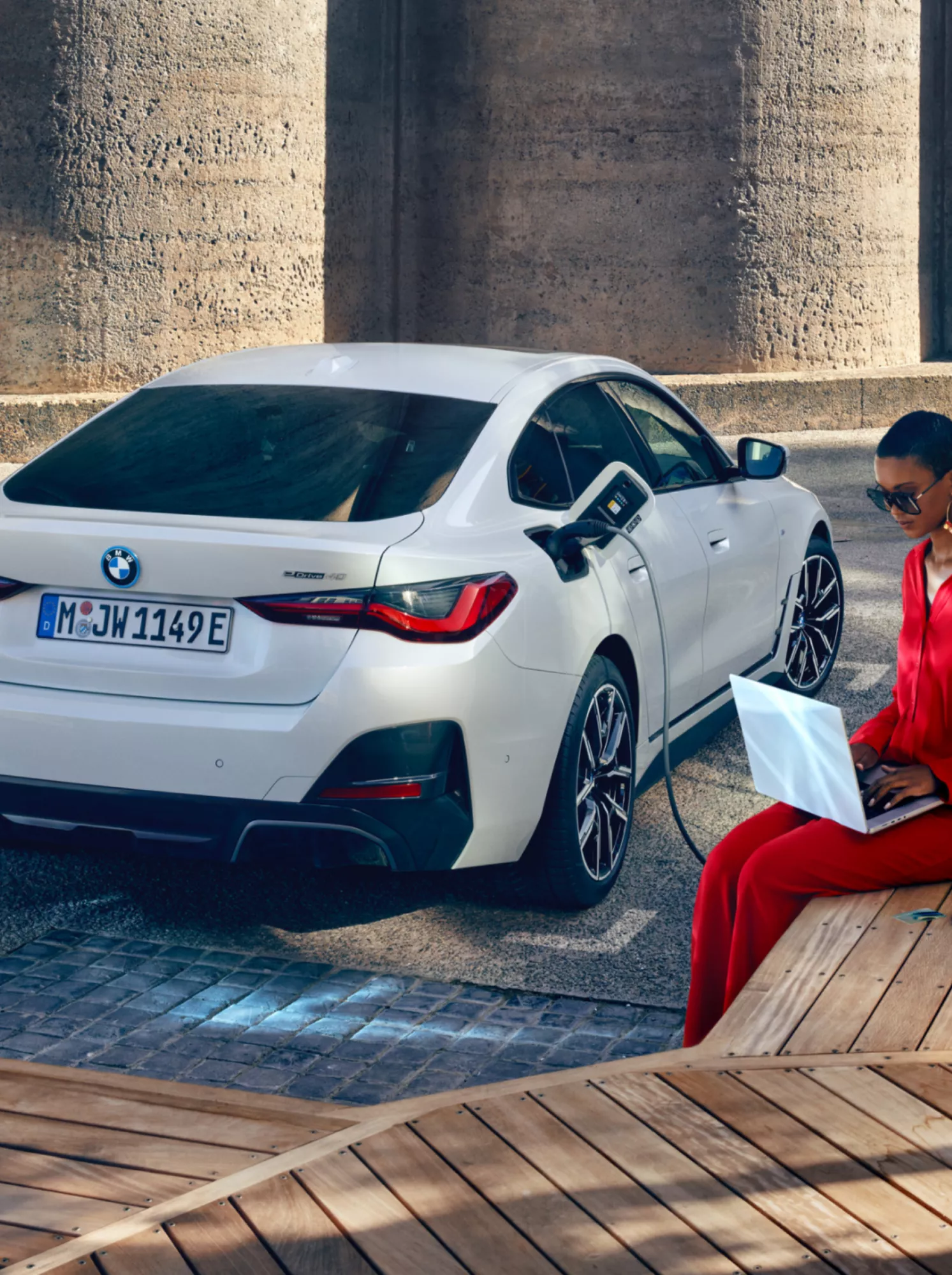 Un’auto BMW è in ricarica presso una colonnina. Una donna in tailleur pantalone è seduta comodamente accanto e lavora con un laptop