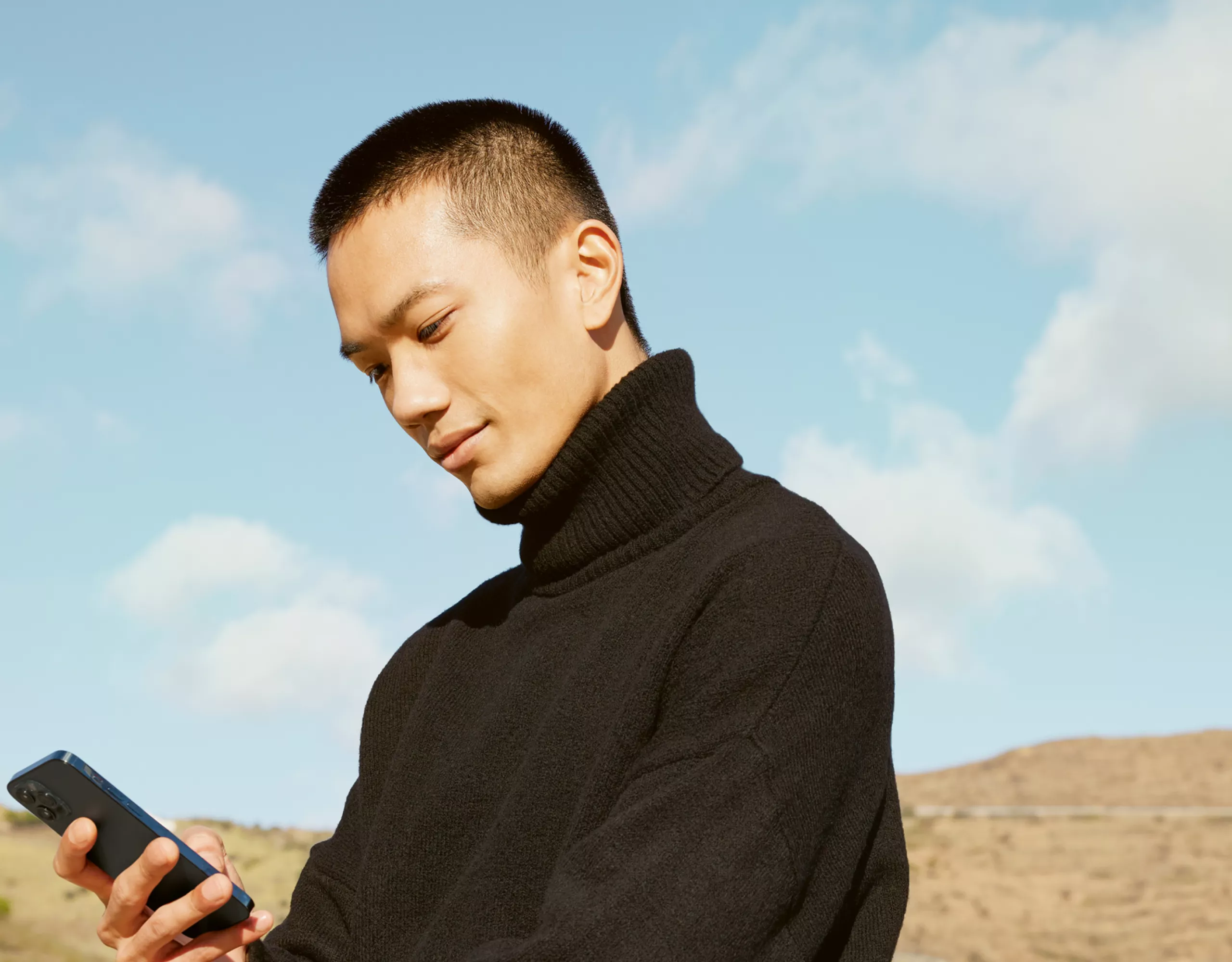 Ein Mann in schwarzem Rollkragenpullover lehnt am Heck seines BMW und schaut in sein Smartphone.