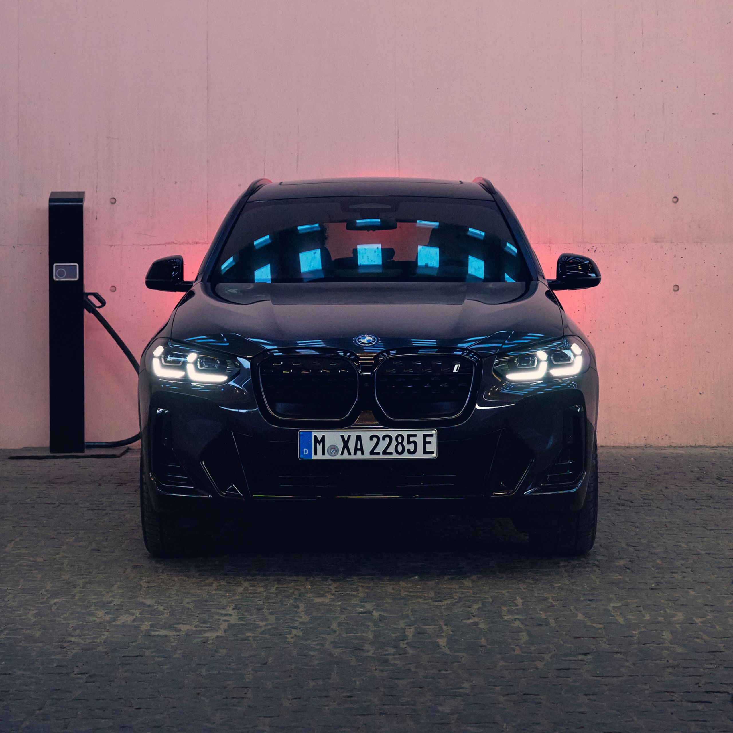 Die Frontansicht eines schwarzen BMW. Der BMW steht neben einer Ladesäule.