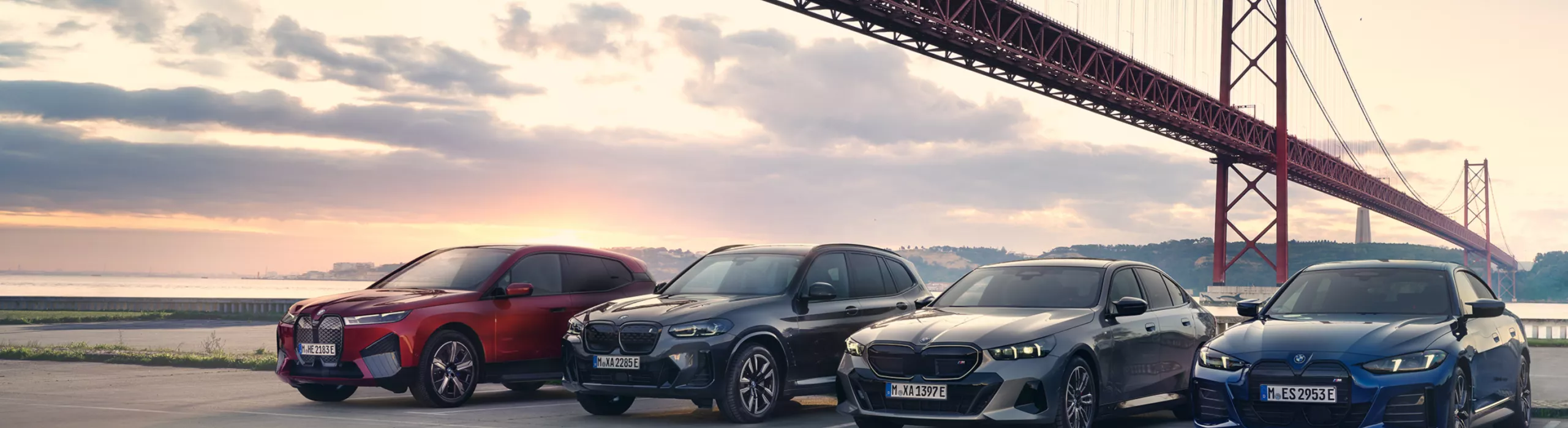 Vier BMW in unterschiedlichen Farben stehen unter einer Hängebrücke mit Blick aufs Meer.