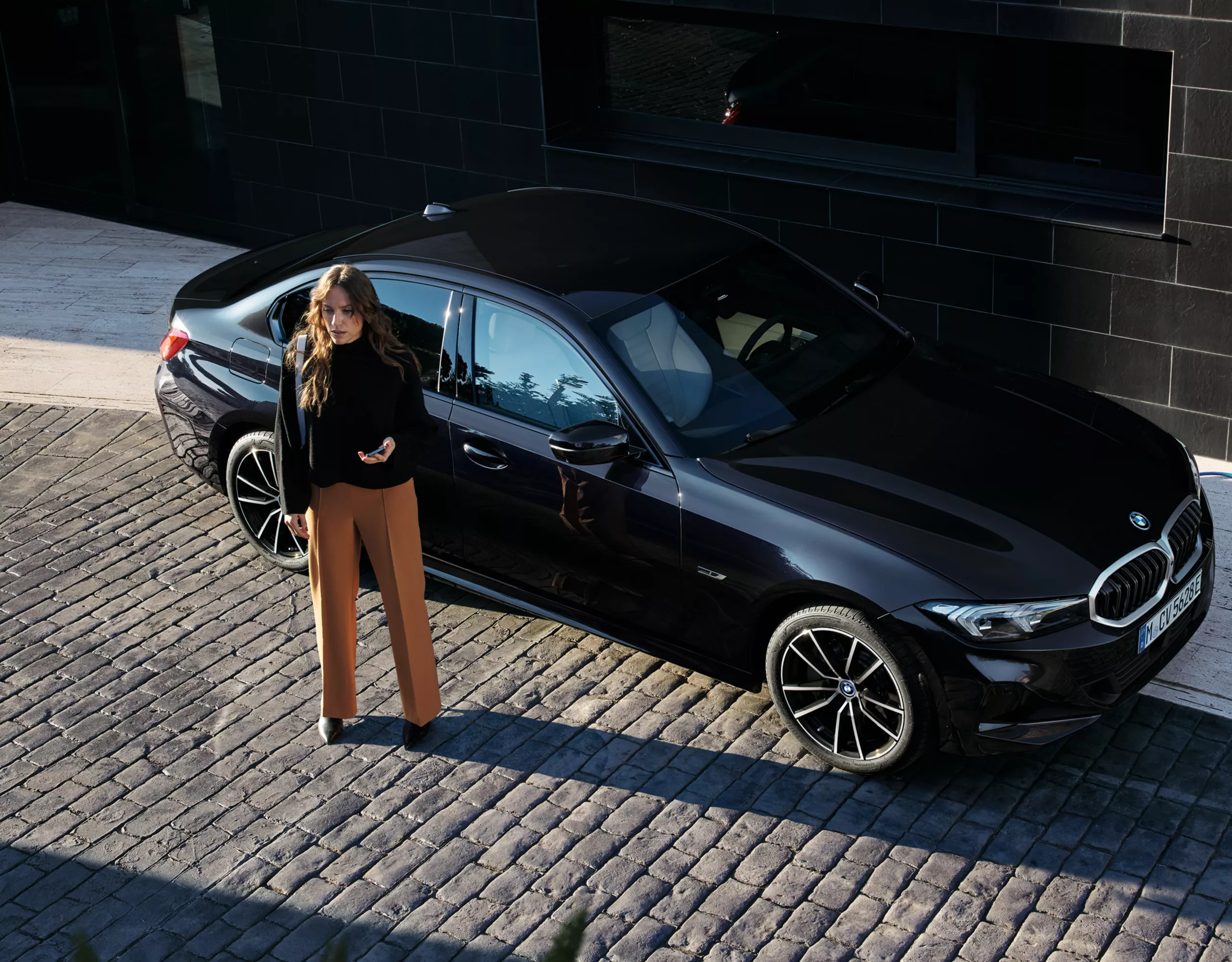 充電中のBMWプラグイン・ハイブリッド・モデルと、その隣に立つスマートフォンを持っている女性