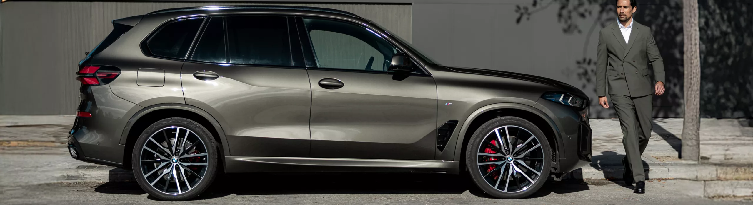 En BMW X5 Plug in Hybrid (PHEV) i Manhattan Metallic står parkerad framför en modern arkitektonisk byggnad. En person står avslappnat bredvid förarsidan av en lyxig SUV och framhäver dess premiumdesign, sportiga proportioner och eleganta metallic-finish