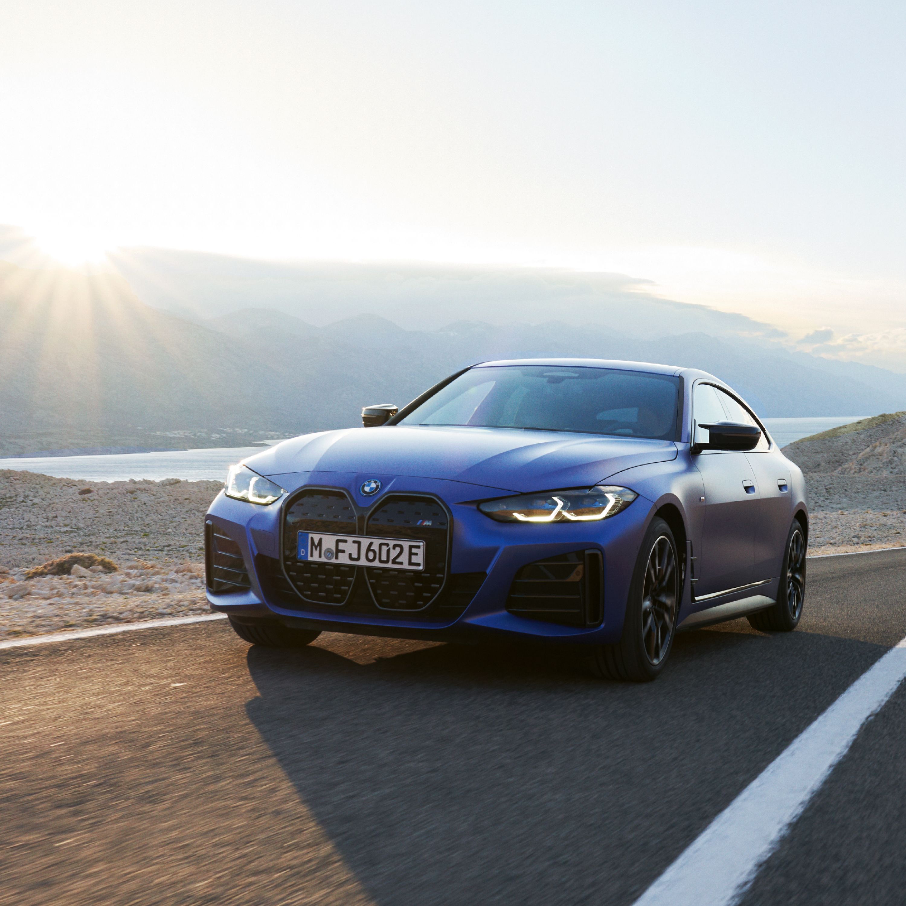 BMW i4 M50 Gran Coupé in Tansanitblau Metallic auf einer Bergstraße in der Nähe eines Sees mit Sonnenaufgang im Hintergrund