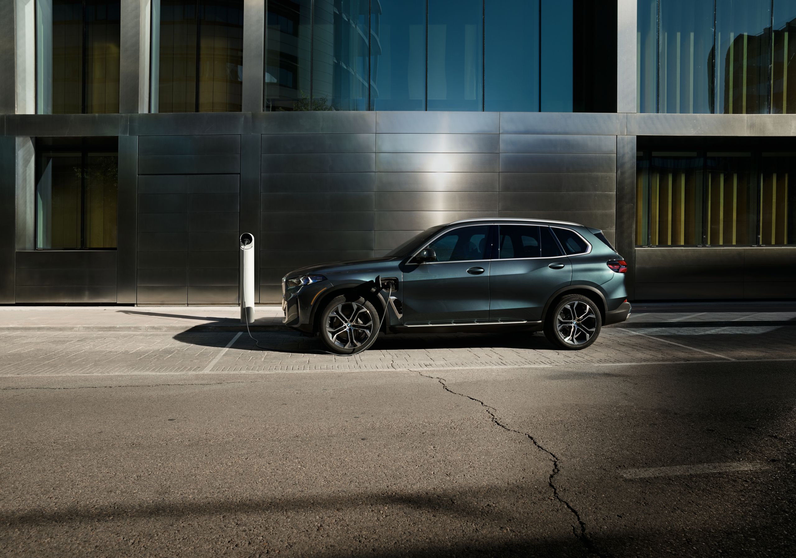 Side profile view of the BMW X5 xDrive50e charging at an outdoor charger in an urban landscape