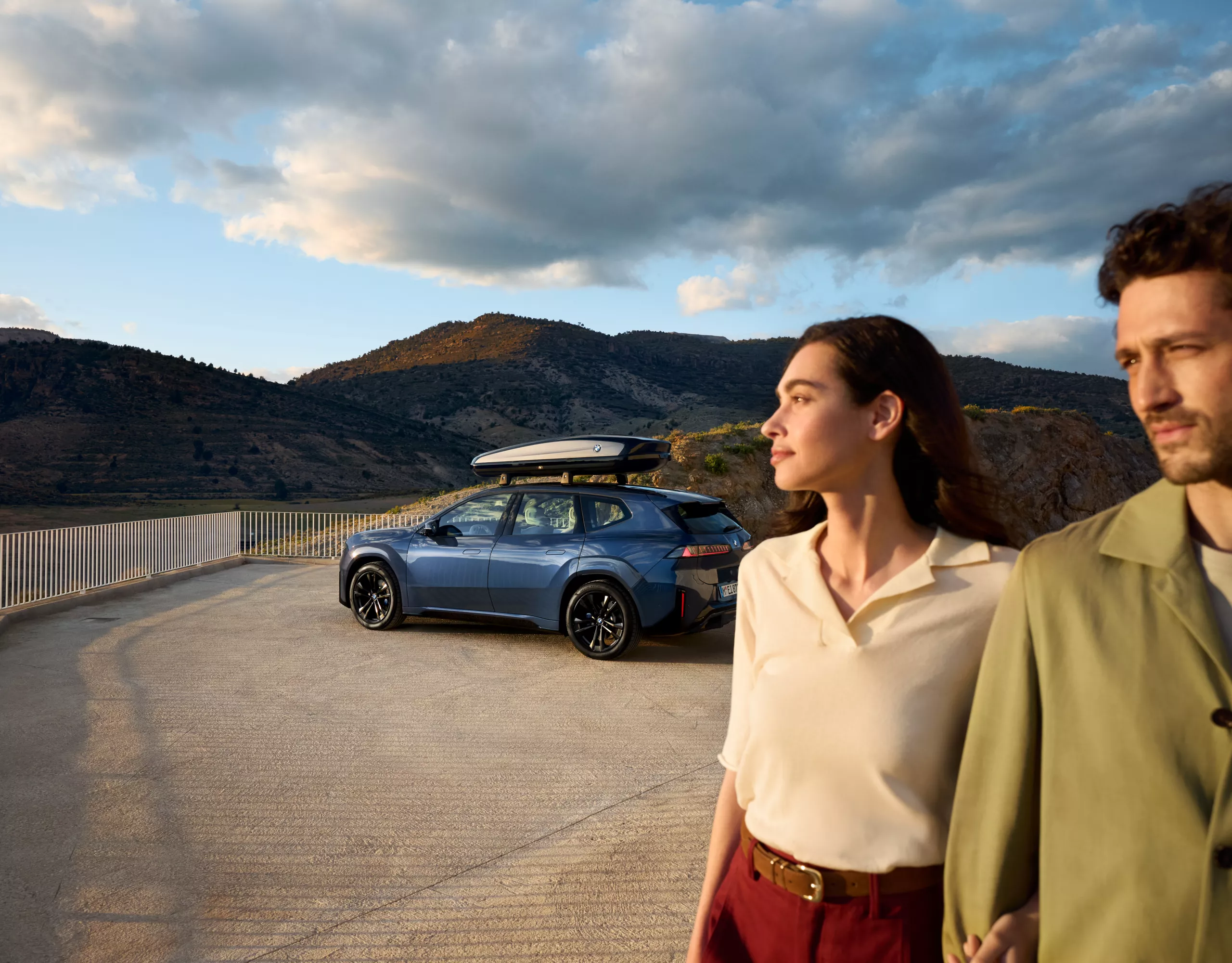 A couple stands in front of a BMW iX3 on a platform against a backdrop of mountains