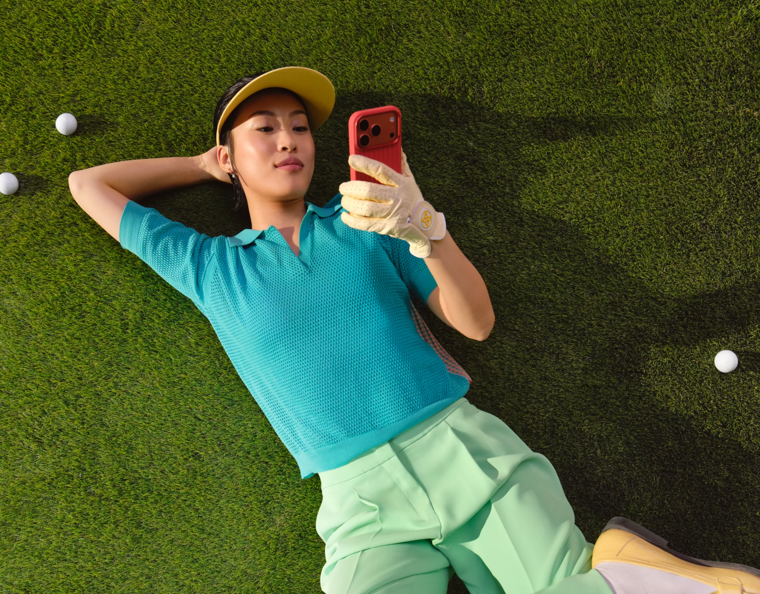 Una donna è sdraiata su un prato con uno smartphone in mano, circondata da palline da golf