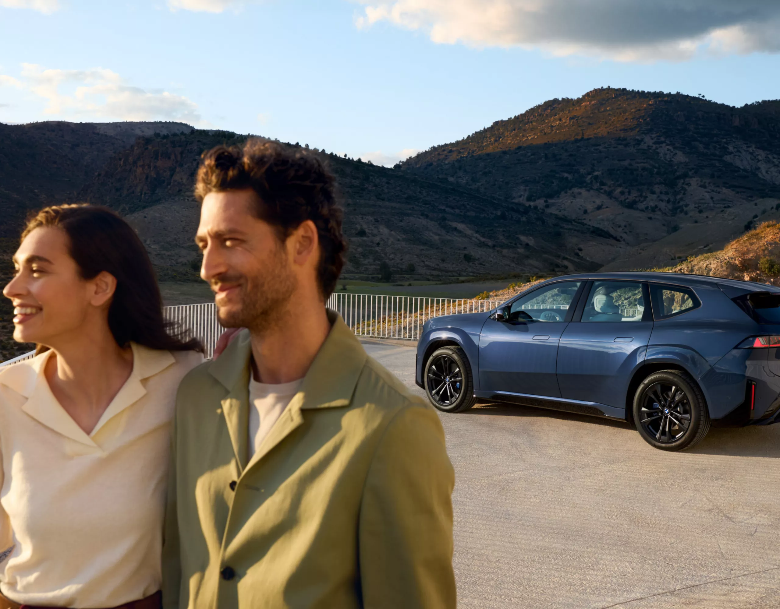 A couple is standing in front of a BMW iX3 on a platform against a backdrop of mountains