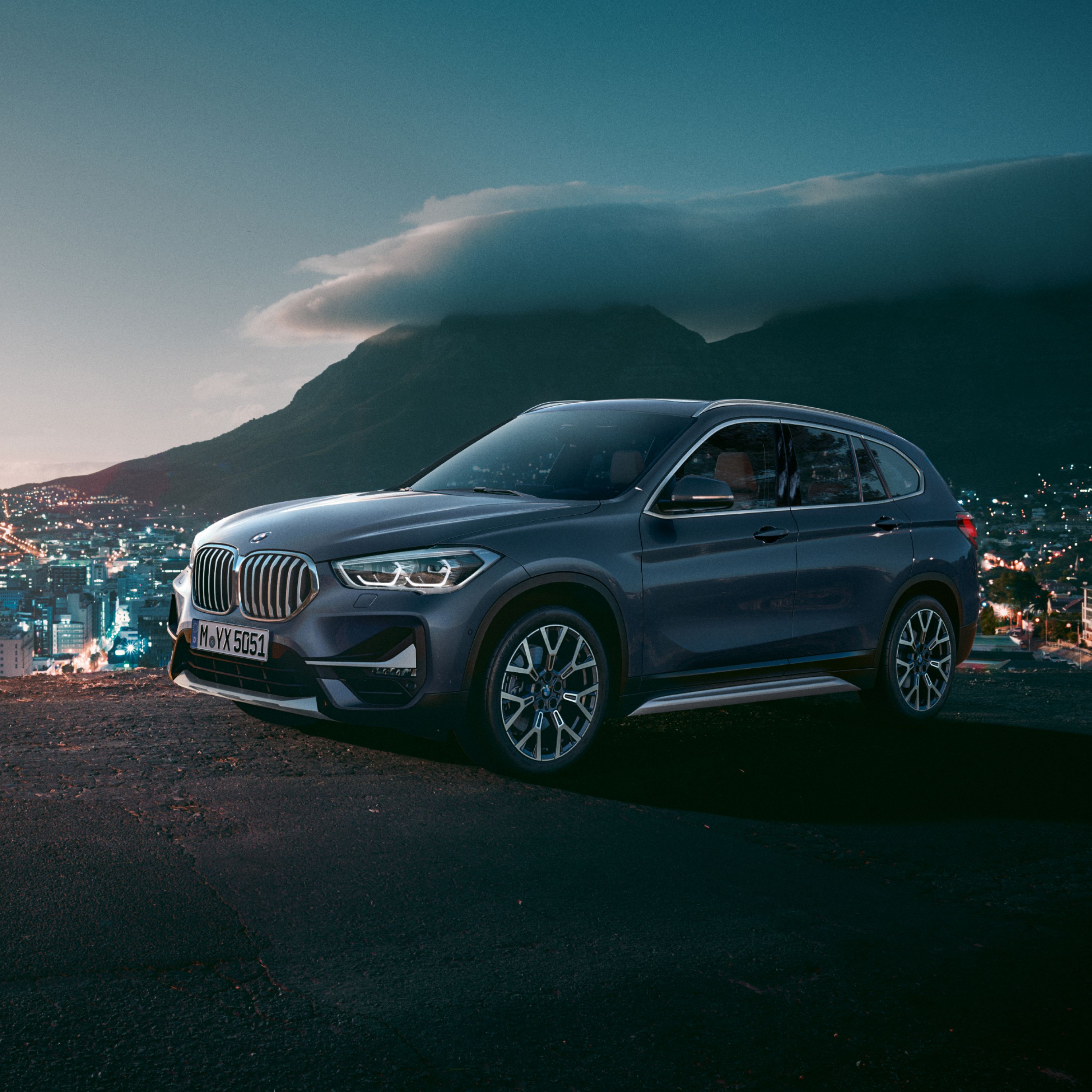 BMW X1 F48 SUV auf einer Anhöhe mit Ausblick auf ein Städtepanorama mit Berglandschaft im Hintergrund