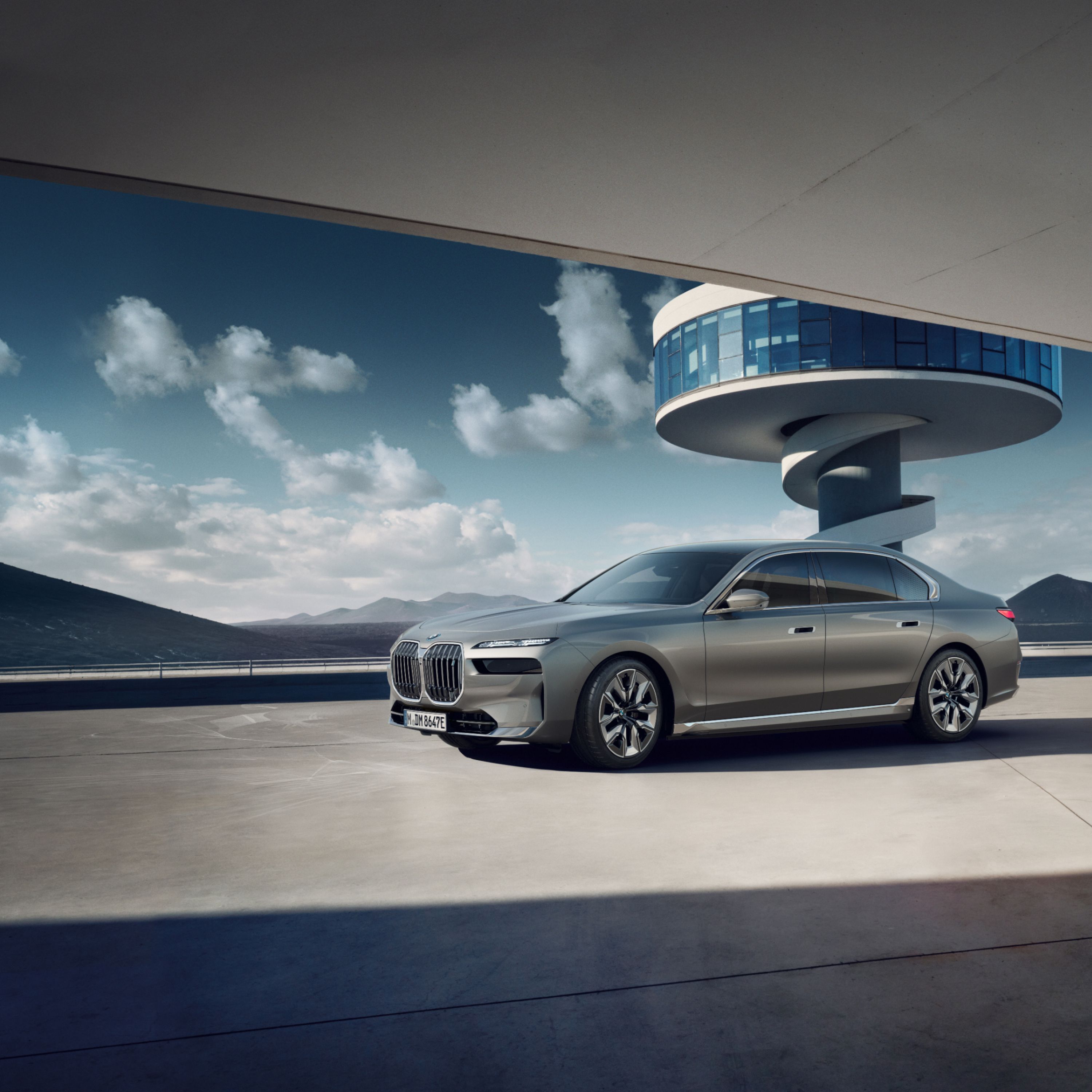 BMW i7 xDrive60 on a tarmac in front of a tower with a spiral staircase and a mountain panorama behind it. 