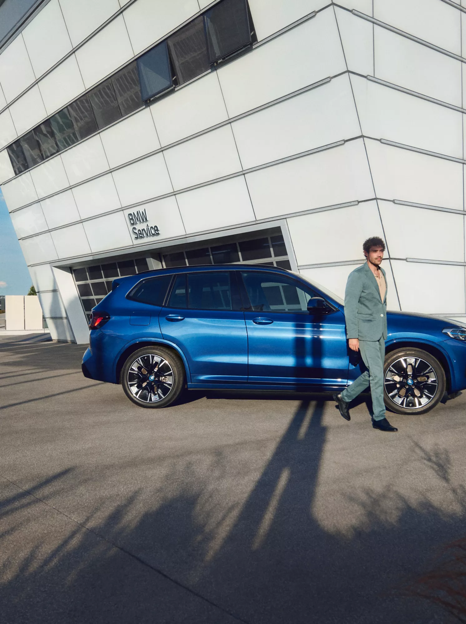 Мъж в сиво-зелен костюм и жена с жълт панталон вървят около син BMW SUV, паркиран пред бяла сграда на BMW Service с модерна архитектура