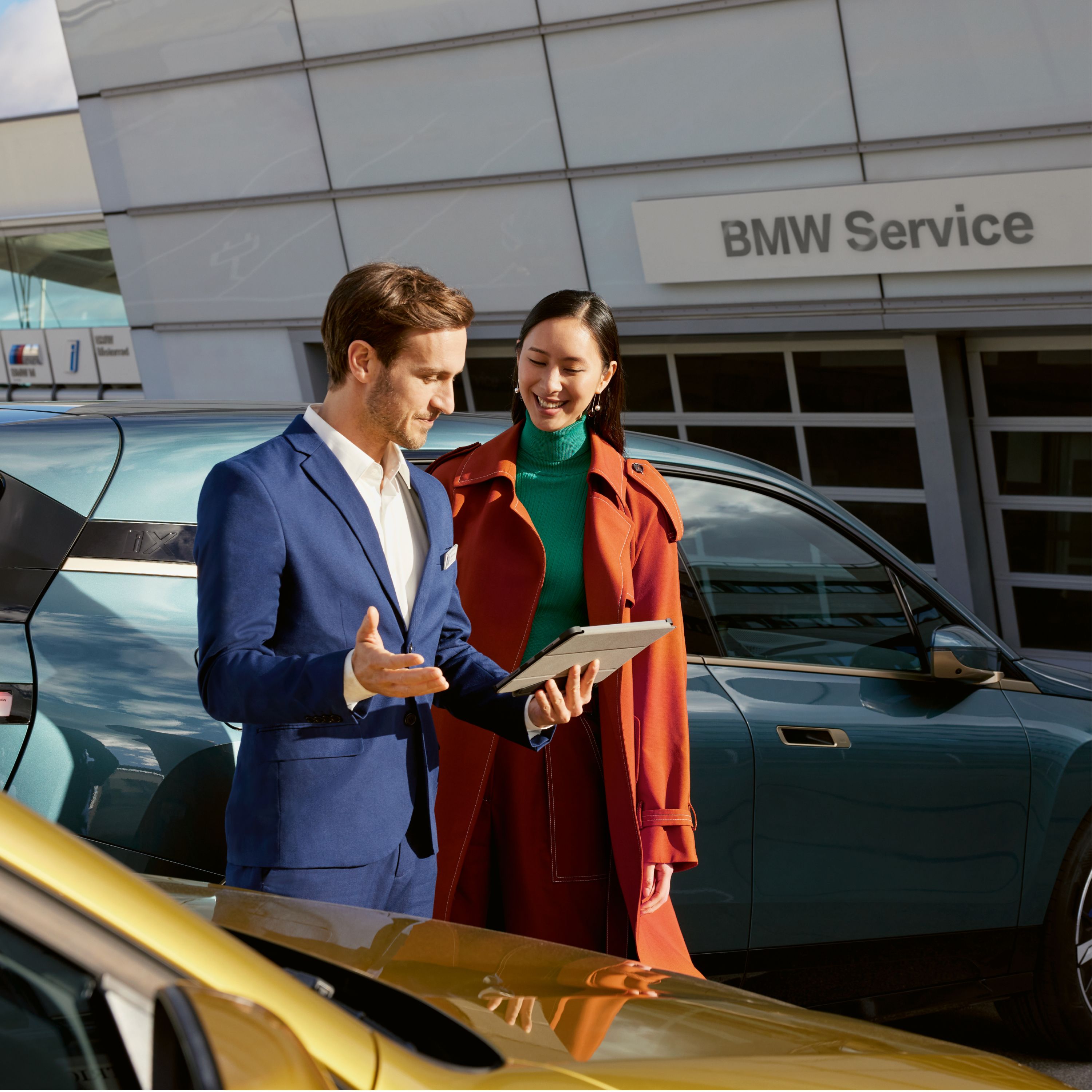 BMW Service, moški in ženska stojita pred BMW iX pri servisnem partnerju BMW