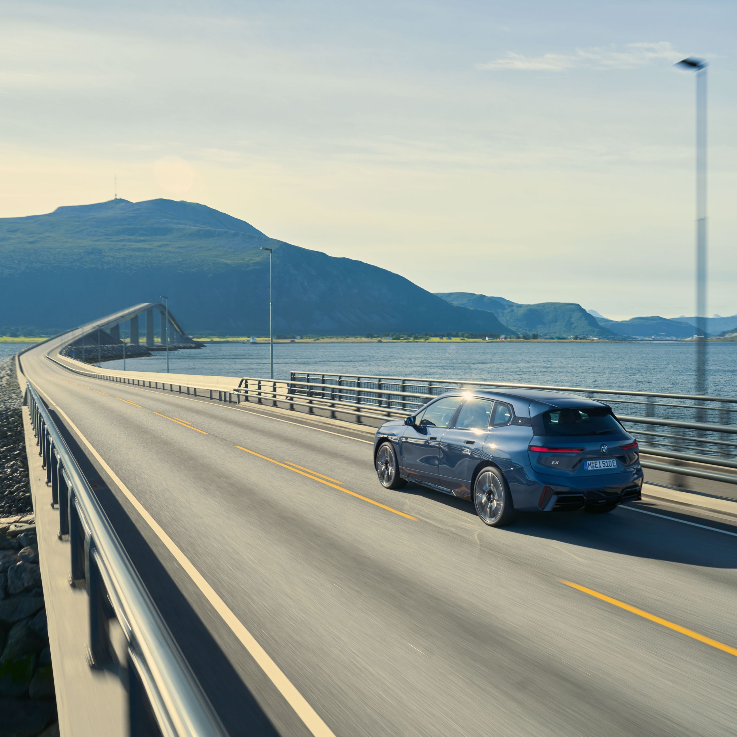 En BMW iX som kör över en bro med vatten och berg i bakgrunden.