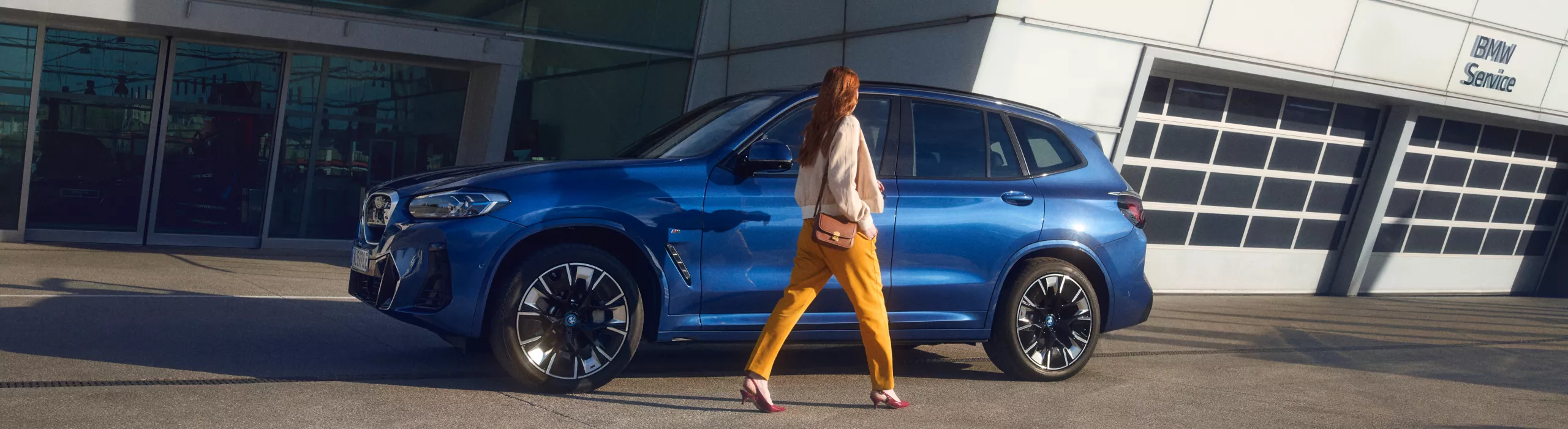 Жена с жълт панталон и светъл пуловер върви към син автомобил BMW пред модерен сервизен център на BMW