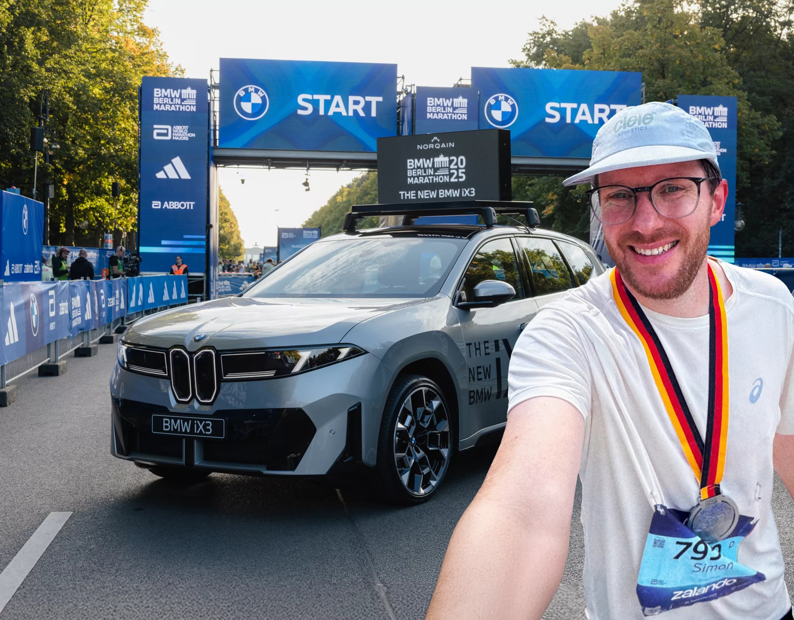  Simon mit Medaille des BMW Berlin Marathon um den Hals macht ein Selfie von sich im Startbereich des Marathons. Hinter ihm steht der neue BMW iX3 in Grau, weiter im Hintergrund ist das Brandenburger Tor zu sehen.