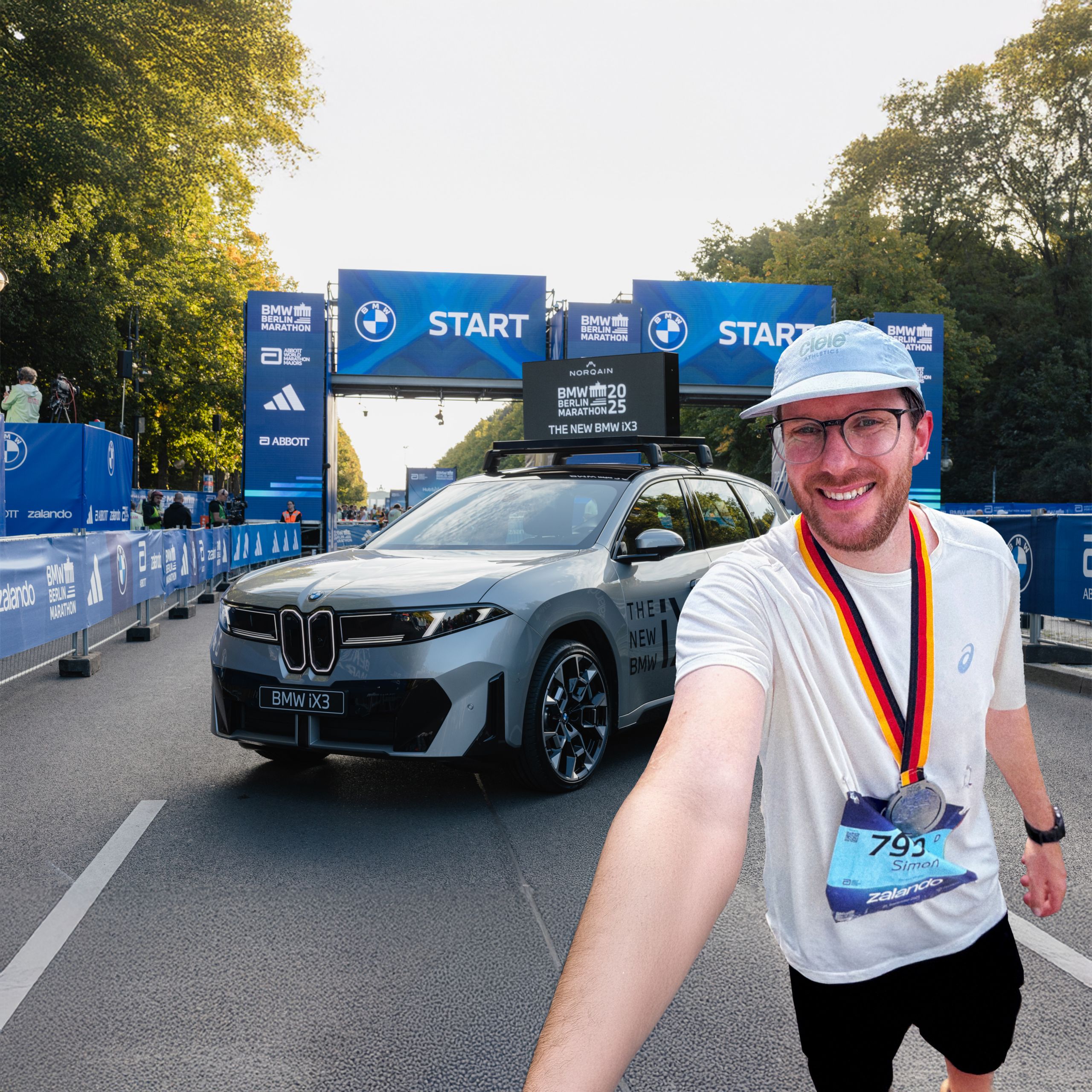  Simon mit Medaille des BMW Berlin Marathon um den Hals macht ein Selfie von sich im Startbereich des Marathons. Hinter ihm steht der neue BMW iX3 in Grau, weiter im Hintergrund ist das Brandenburger Tor zu sehen.