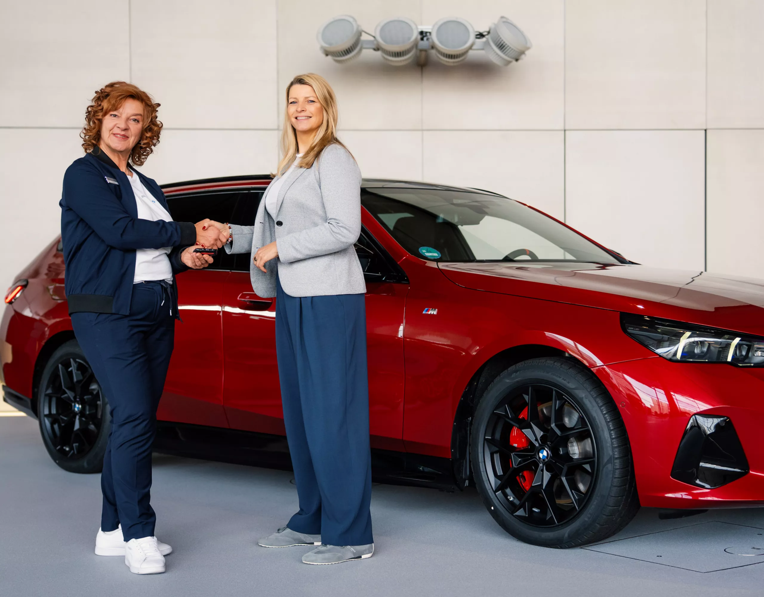  Zwei Frauen stehen vor einem roten BMW i5 M60 xDrive Touring und schütteln sich die Hände. Eine der beiden Frauen ist eine BMW Expertin, die der Kundin den Fahrzeugschlüssel übergibt. Beide schauen in die Kamera.