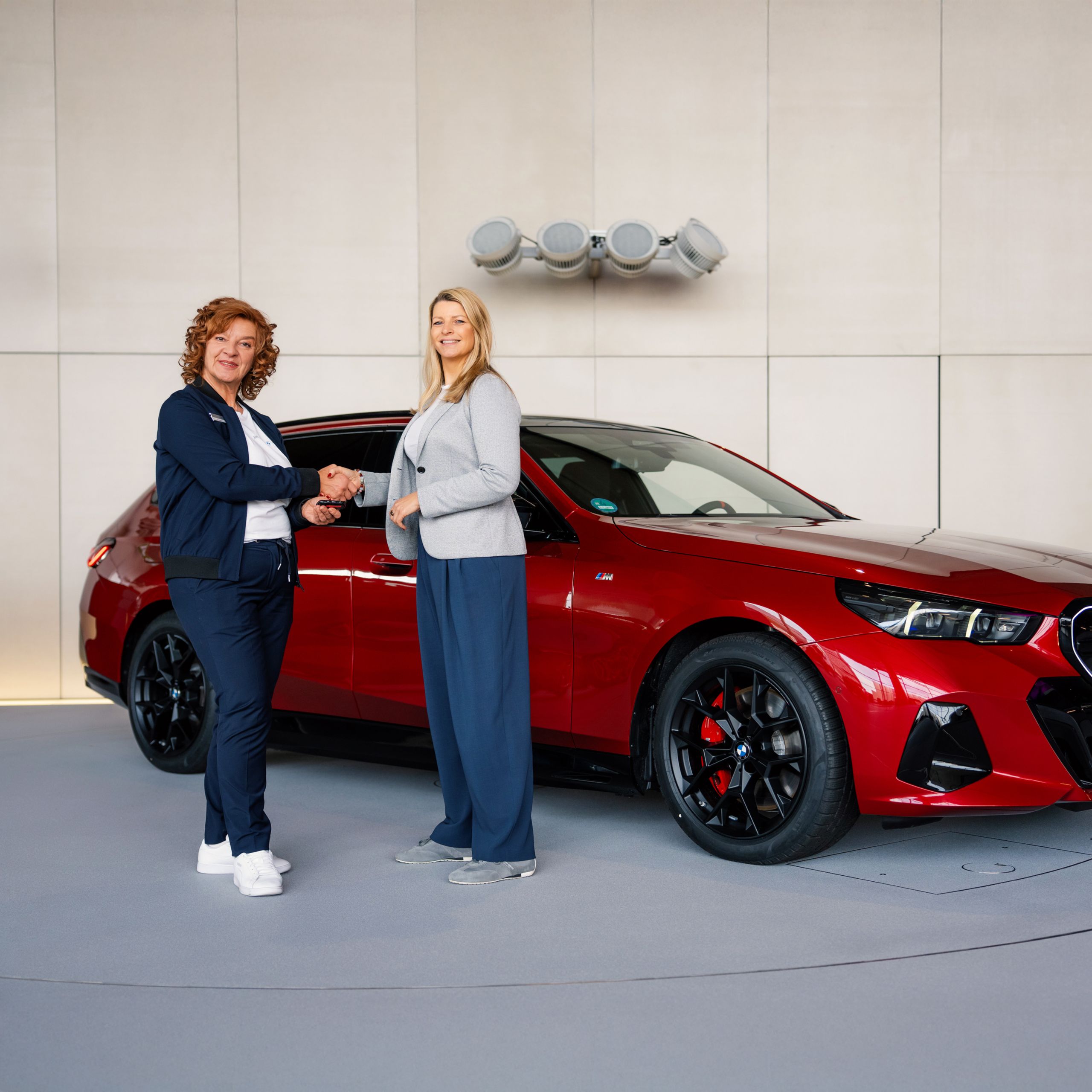  Zwei Frauen stehen vor einem roten BMW i5 M60 xDrive Touring und schütteln sich die Hände. Eine der beiden Frauen ist eine BMW Expertin, die der Kundin den Fahrzeugschlüssel übergibt. Beide schauen in die Kamera.