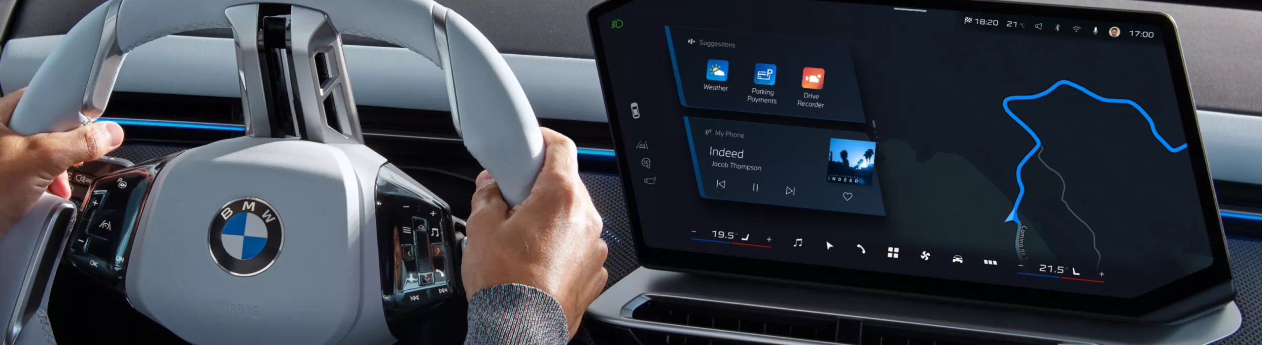 A person is driving a BMW with both hands on the steering wheel and BMW Maps navigation is displayed on the central display