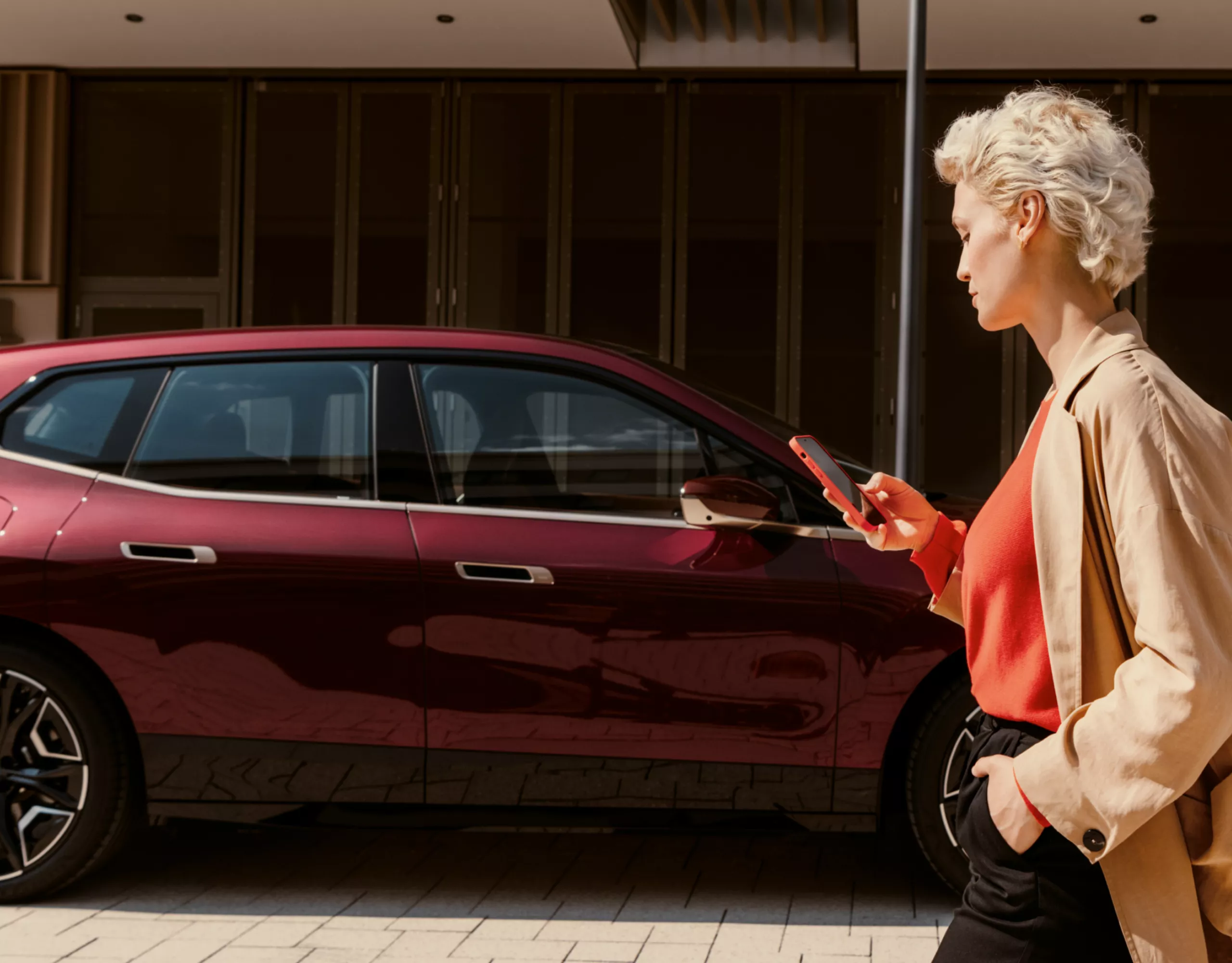 A woman looks at her smartphone while walking with a parked BMW in front of a building in the background