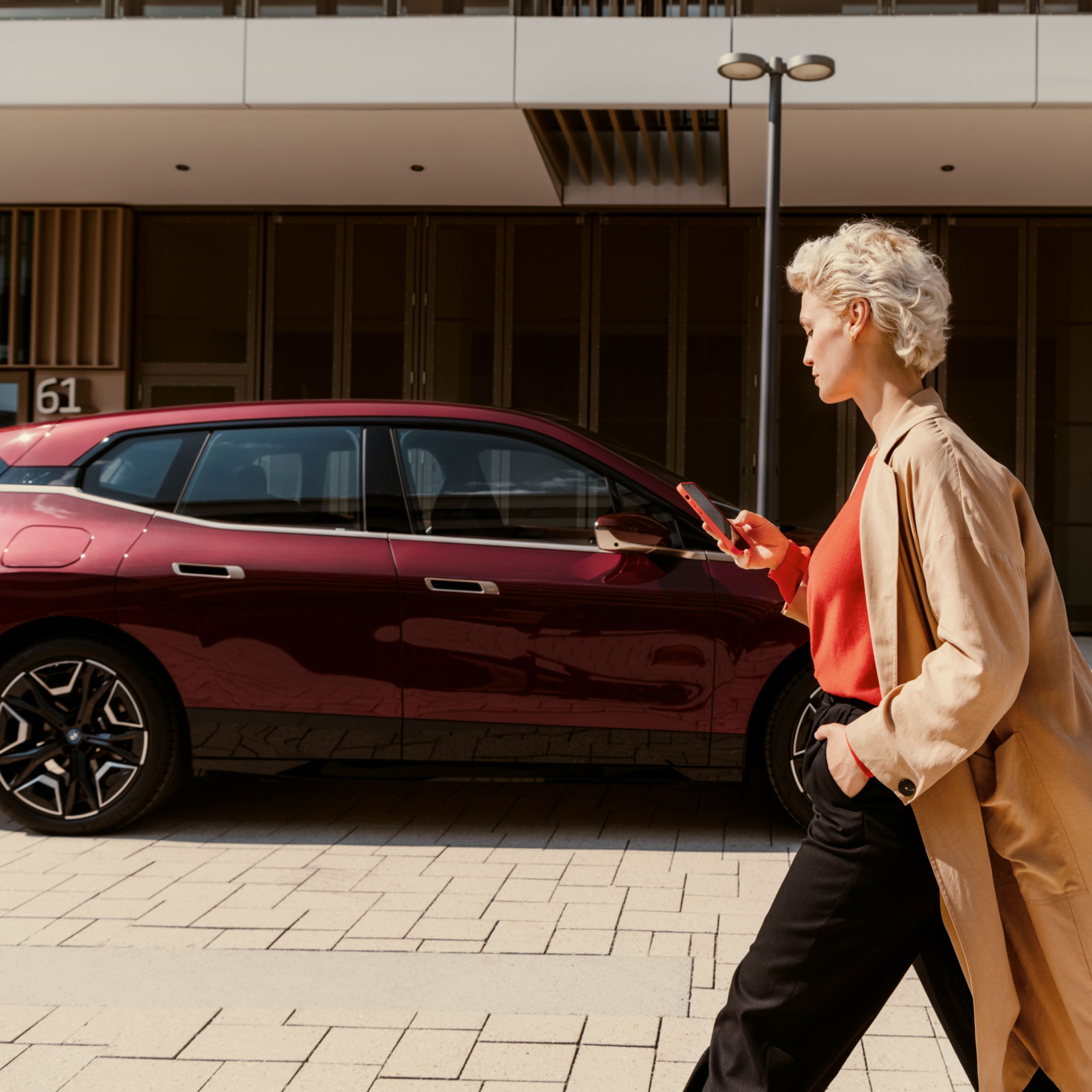 A woman looks at her smartphone while walking with a parked BMW in front of a building in the background