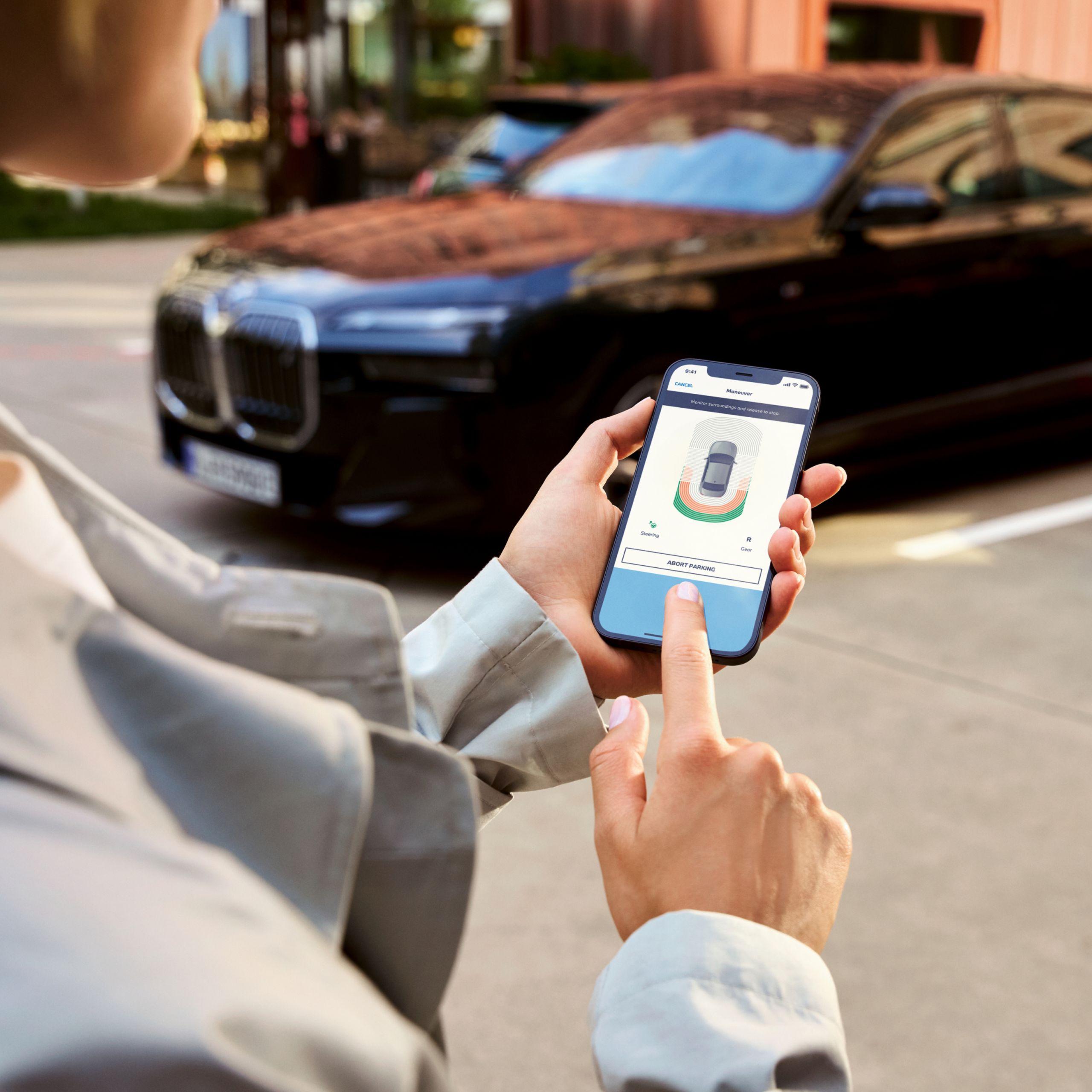 A smartphone with the My BMW App is being used to park a BMW visible in the background
