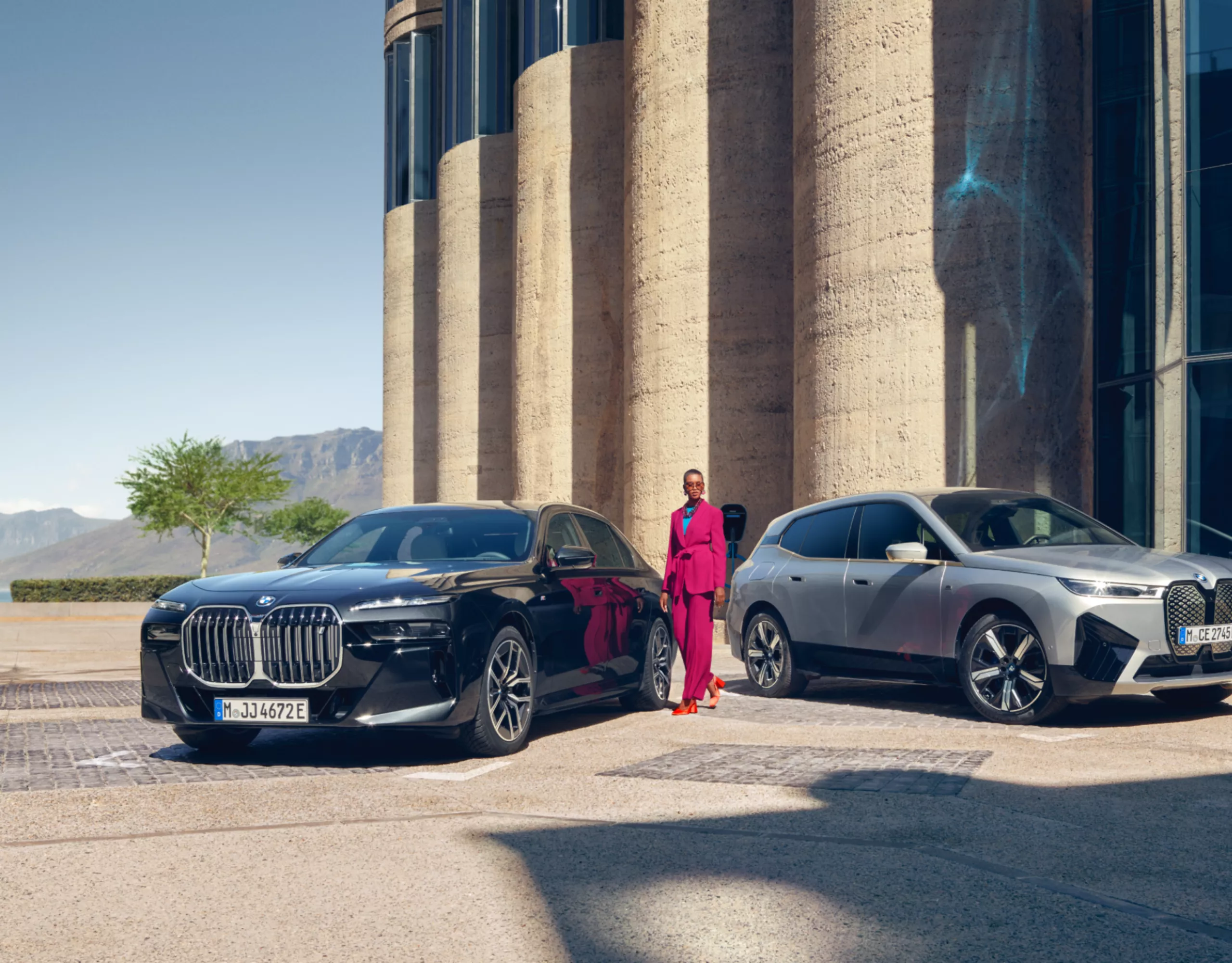 A woman is standing between two fully electric BMW vehicles in front of a building