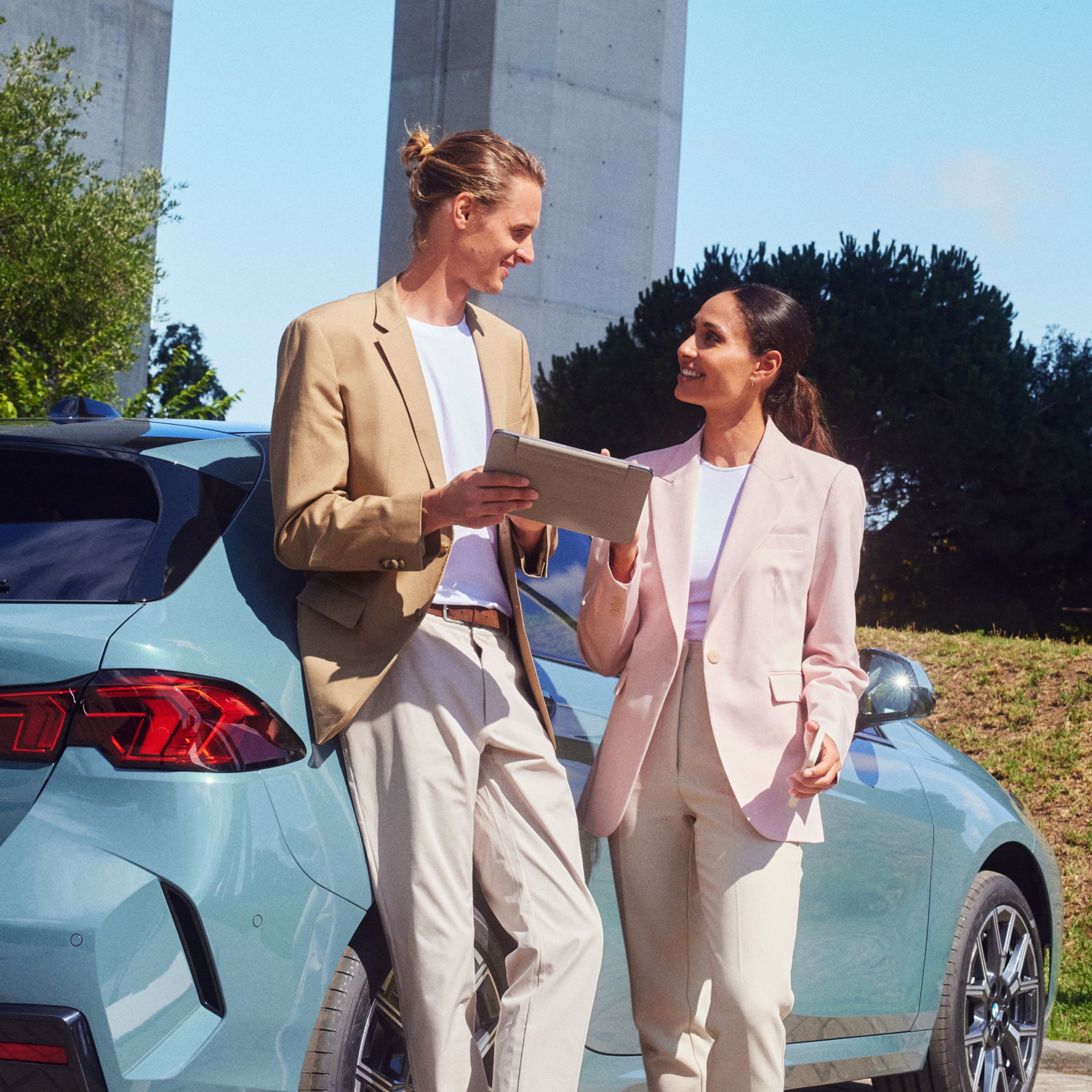 A person with a clipboard is leaning against a BMW and talking to another person