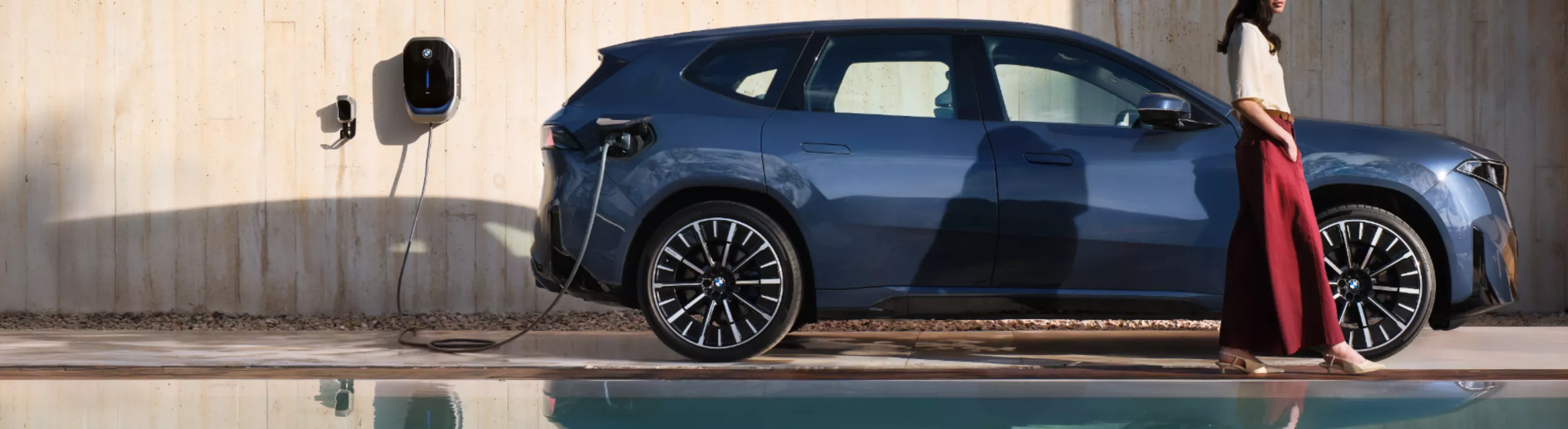 A person walks away from a BMW charging at a wallbox, with the scene reflected in an ornamental pond