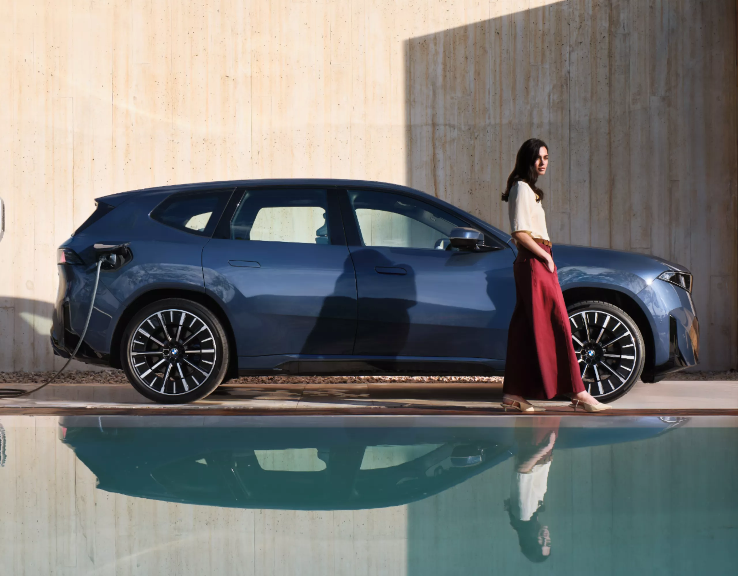 A person walks away from a BMW charging at a wallbox, with the scene reflected in an ornamental pond
