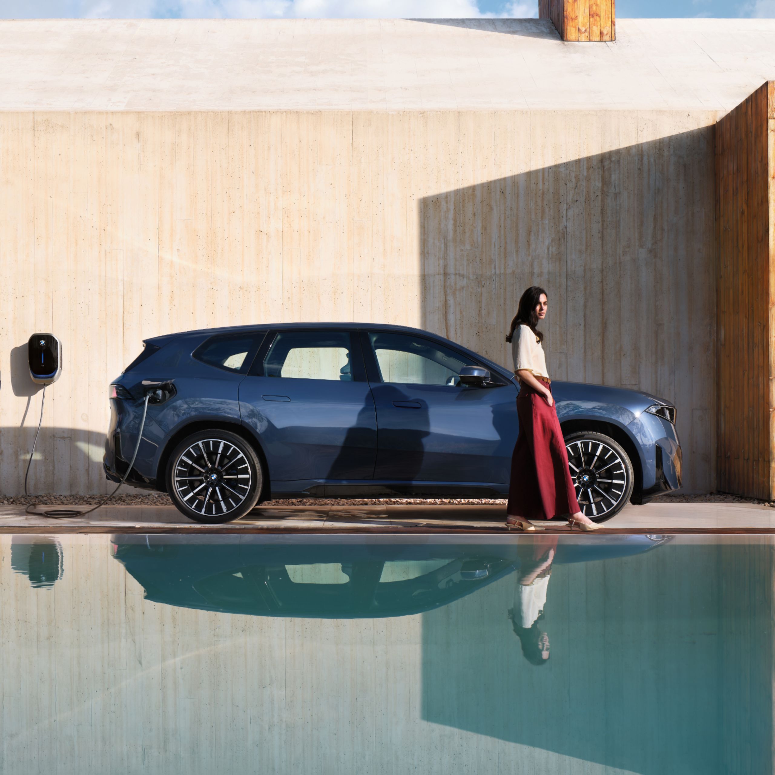 A person walks away from a BMW charging at a wallbox, with the scene reflected in an ornamental pond