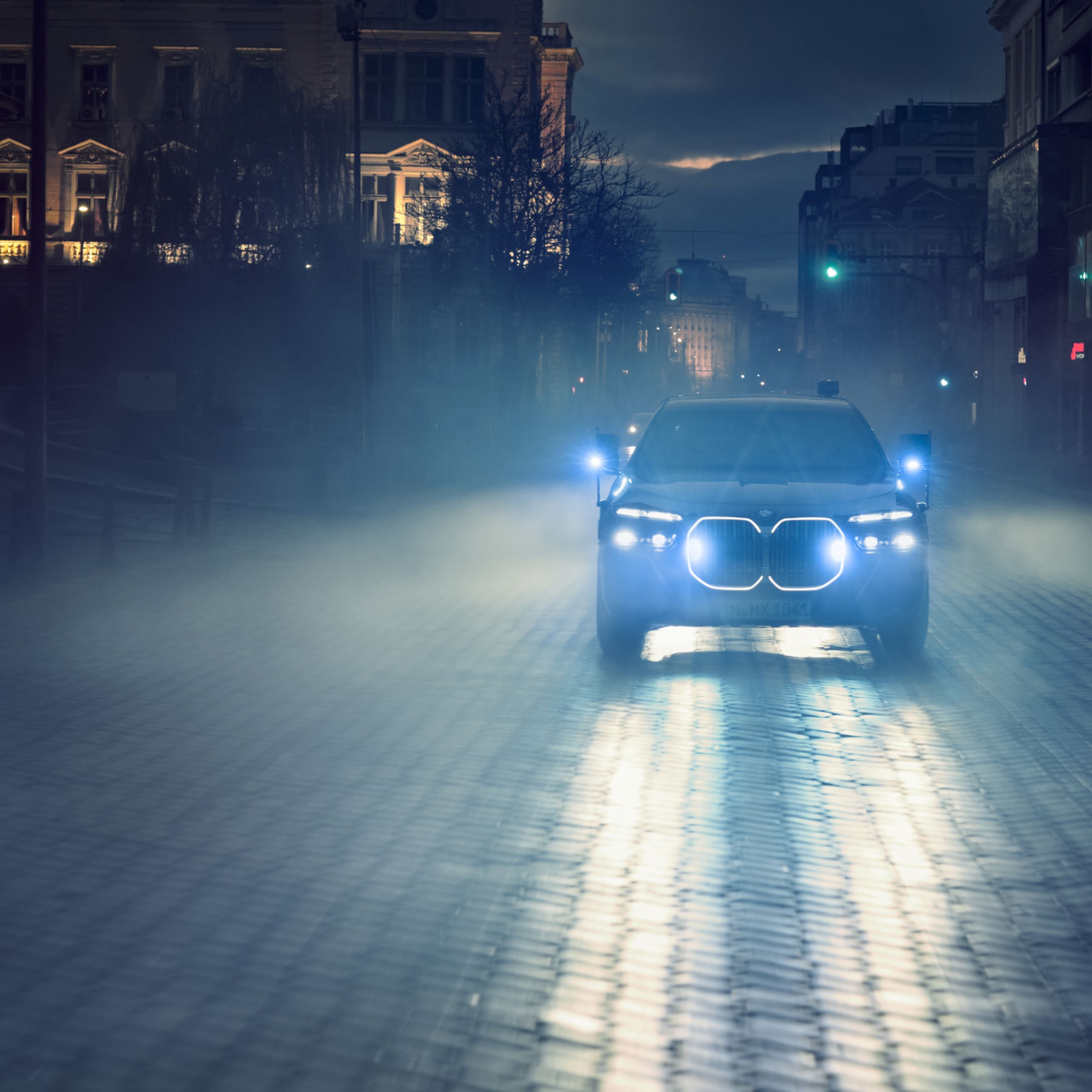 A BMW drives through an old town at night with the front flashing lights switched on and lit up pennants