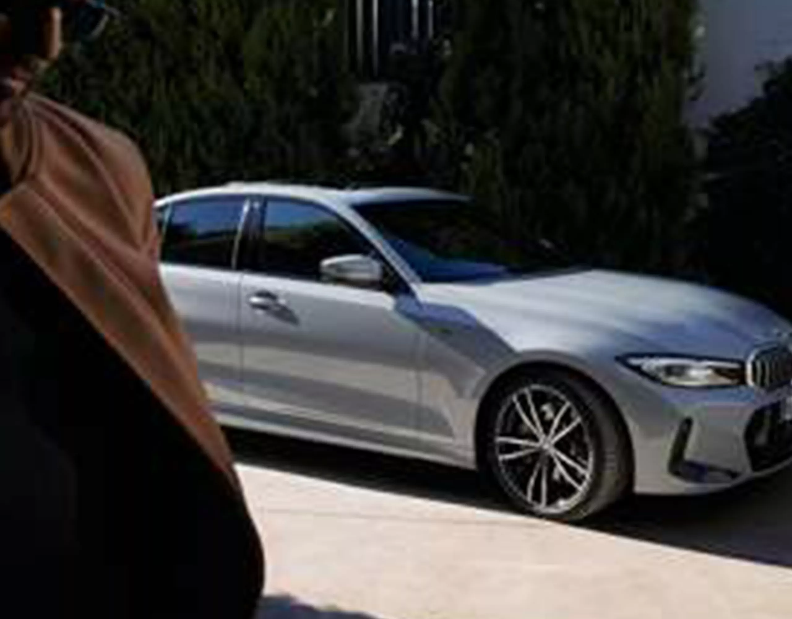 Une femme est assise dans une BMW, la porte ouverte. Un homme est appuyé contre la voiture, avec une maison en arrière-plan.