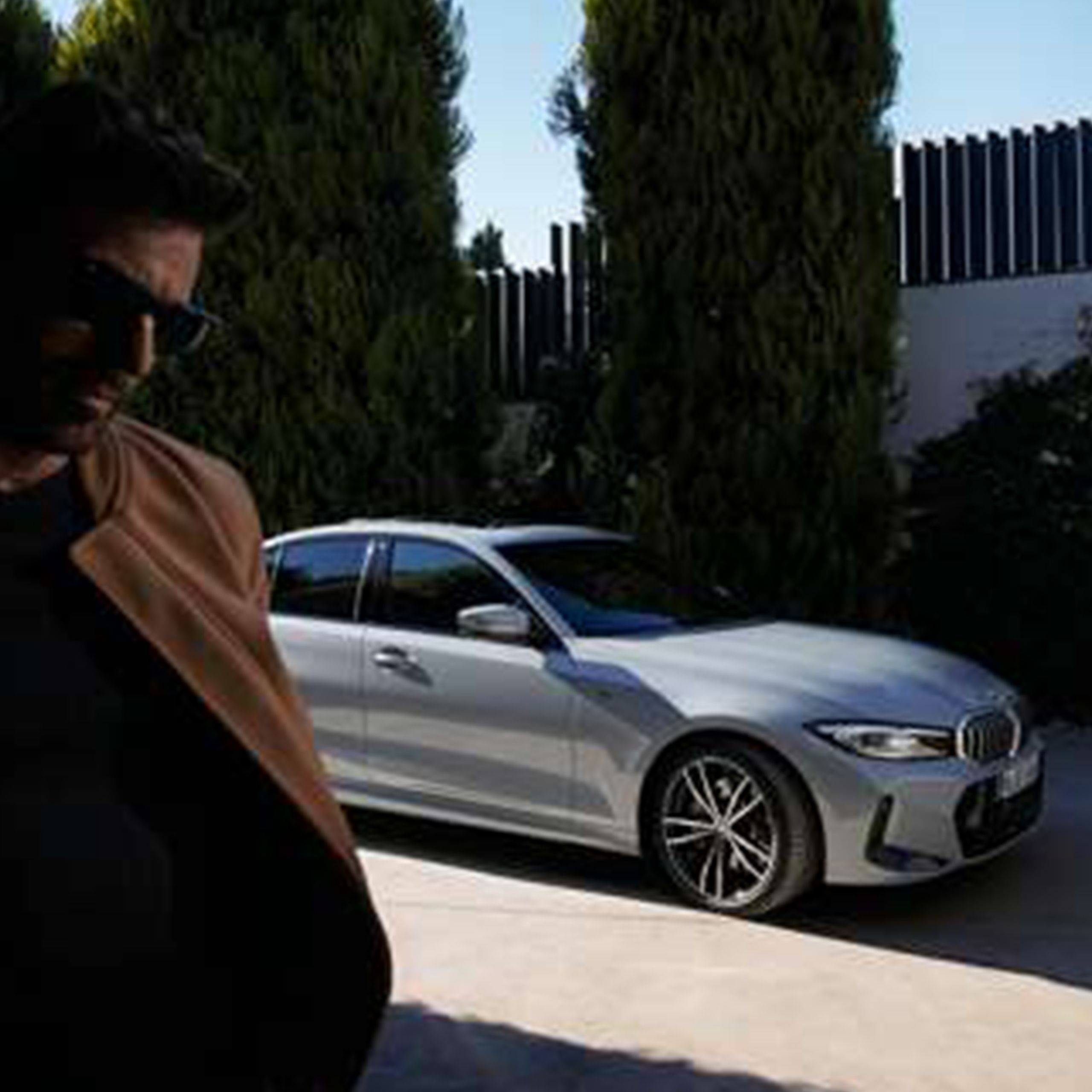 A woman sits in a BMW with the door open. A man leans on the vehicle with a house visible in the background