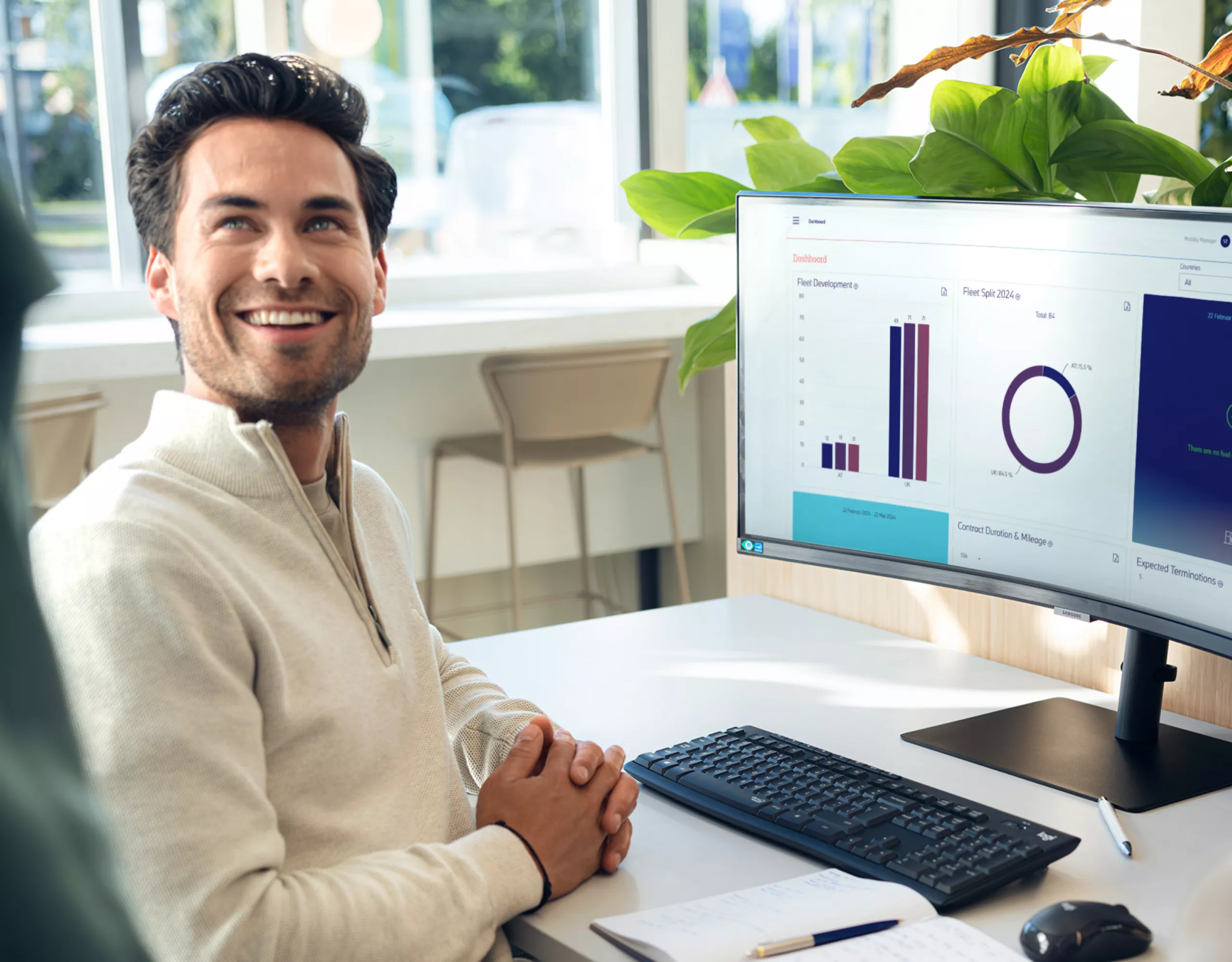 A man sits at a desk with a computer and turns around smiling at another person