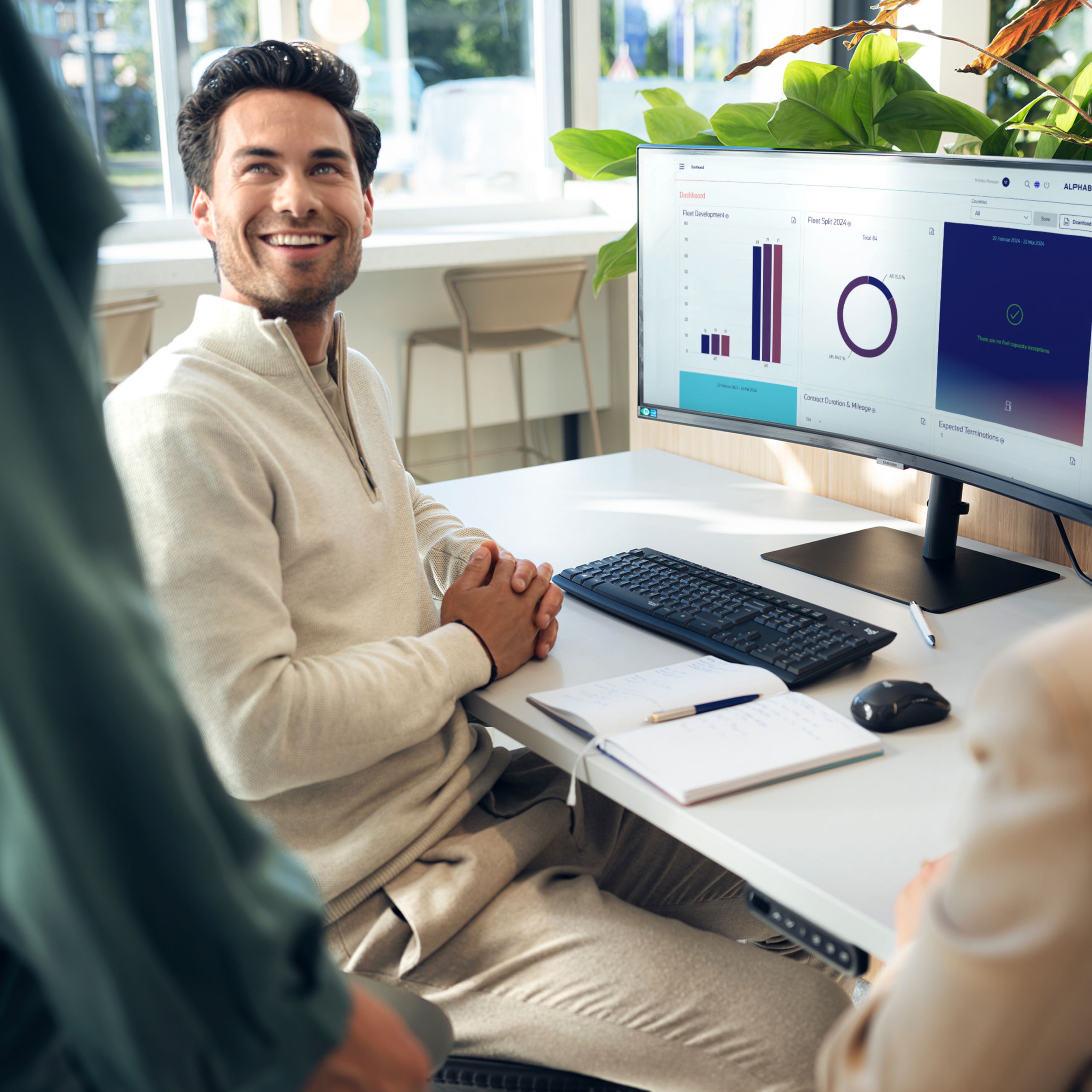 A man sits at a desk with a computer and turns around smiling at another person